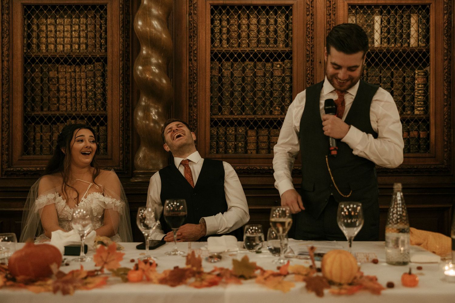 Bride and groom laughing at wedding reception speech in elegant library venue with autumn floral decorations.