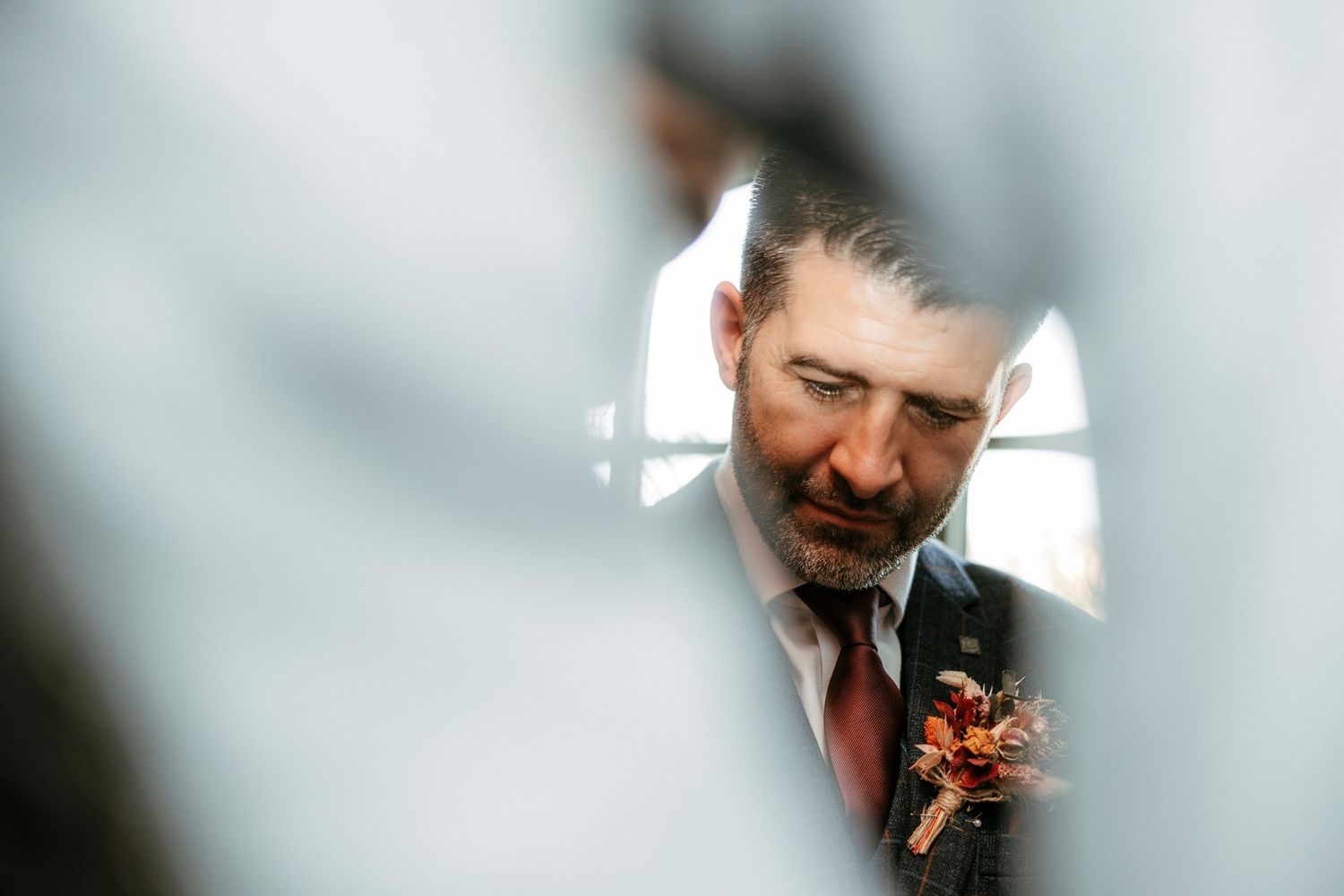 Close-up reflection of groom with beard and boutonniere through delicate fabric.
