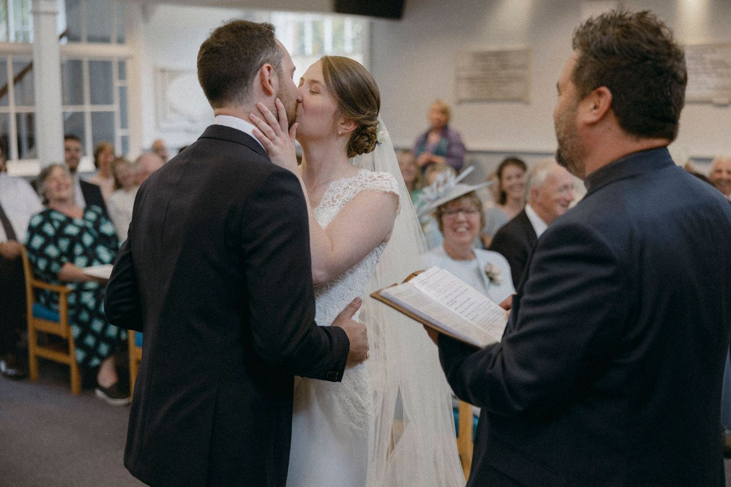 Bride and groom share first kiss at wedding ceremony altar while officiant holds book before seated guests.