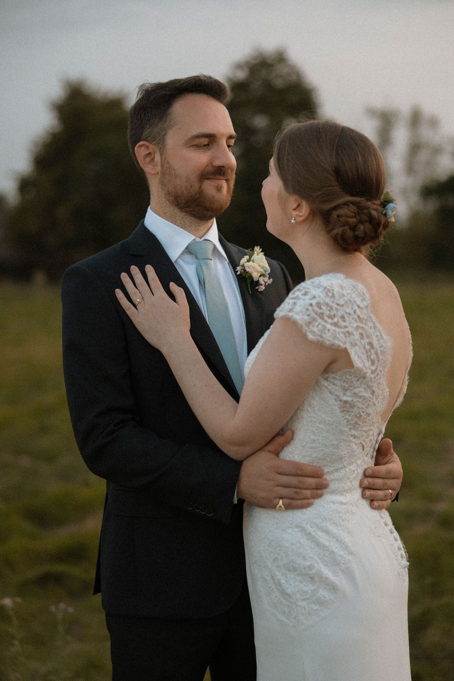 A bride and groom share a tender embrace outdoors, she in lace wedding dress, he in dark suit with blue tie.