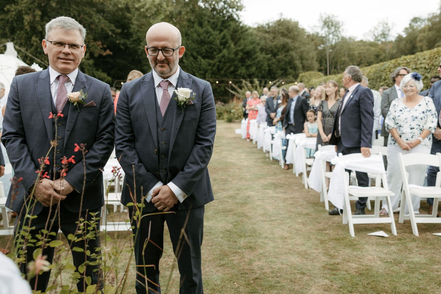 Two grooms in navy suits with boutonnieres stand at outdoor wedding ceremony aisle lined with white chairs.
