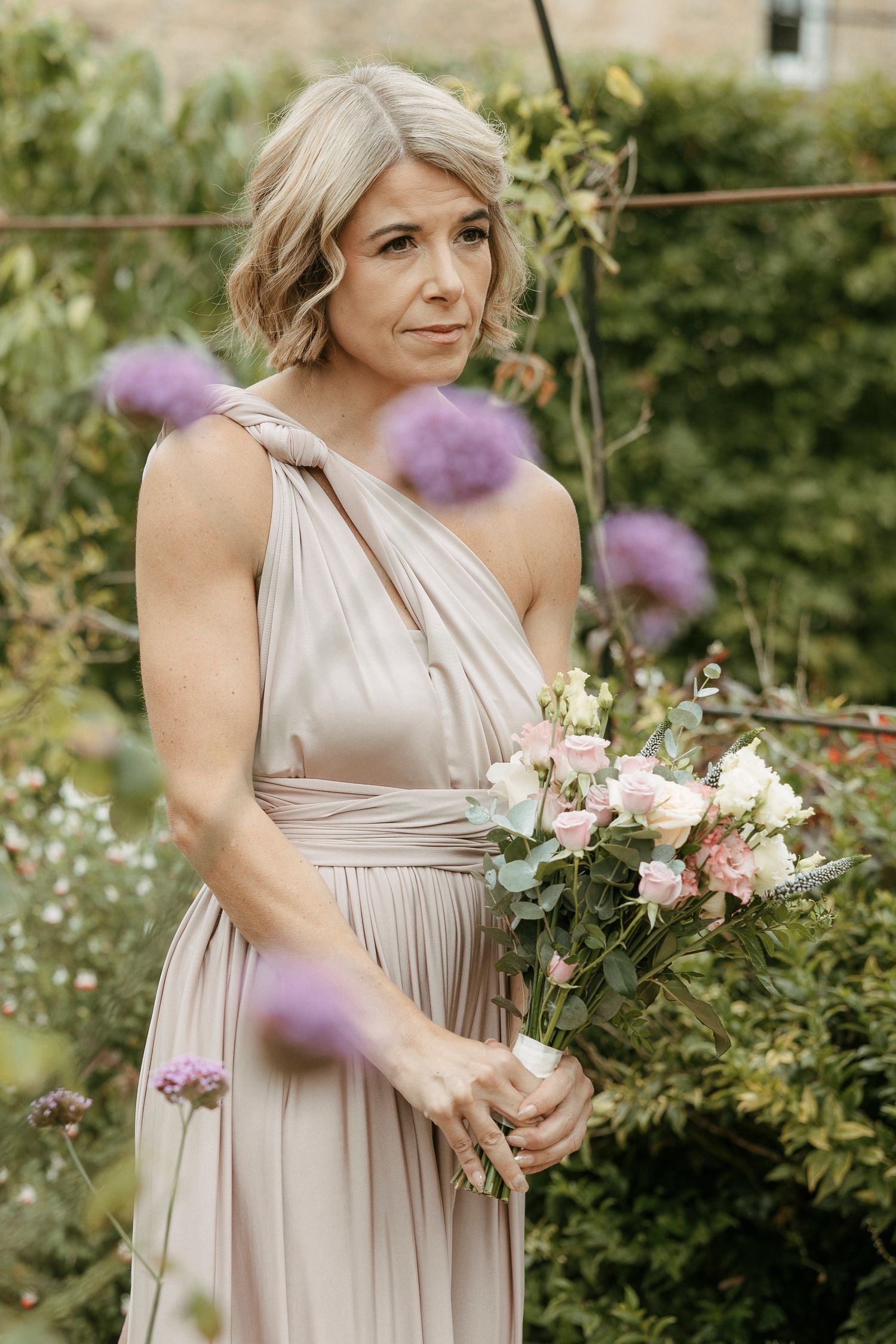 Elegant bridesmaid in champagne one-shoulder gown holding pink floral bouquet in lush garden setting.