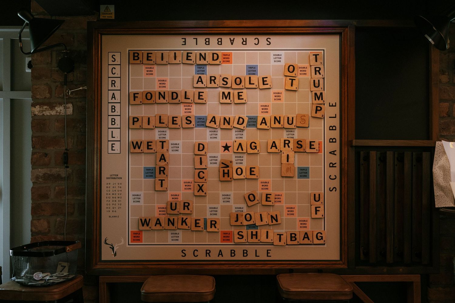 Large Scrabble board display featuring humorous and explicit words arranged in crossword style, mounted on a wooden frame.