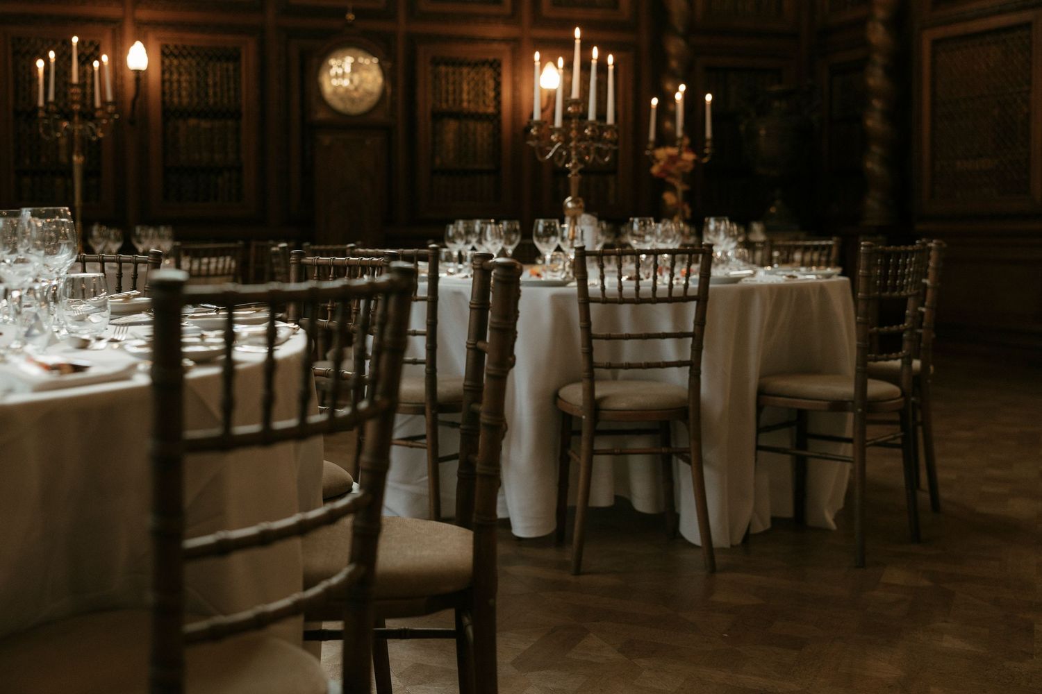 Elegant wedding reception hall with candlelit tables, white linens, and chiavari chairs in a dark wood room.
