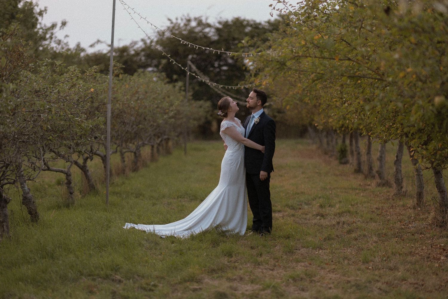 Bride and groom share a romantic moment in a tree-lined orchard path, with string lights overhead.
