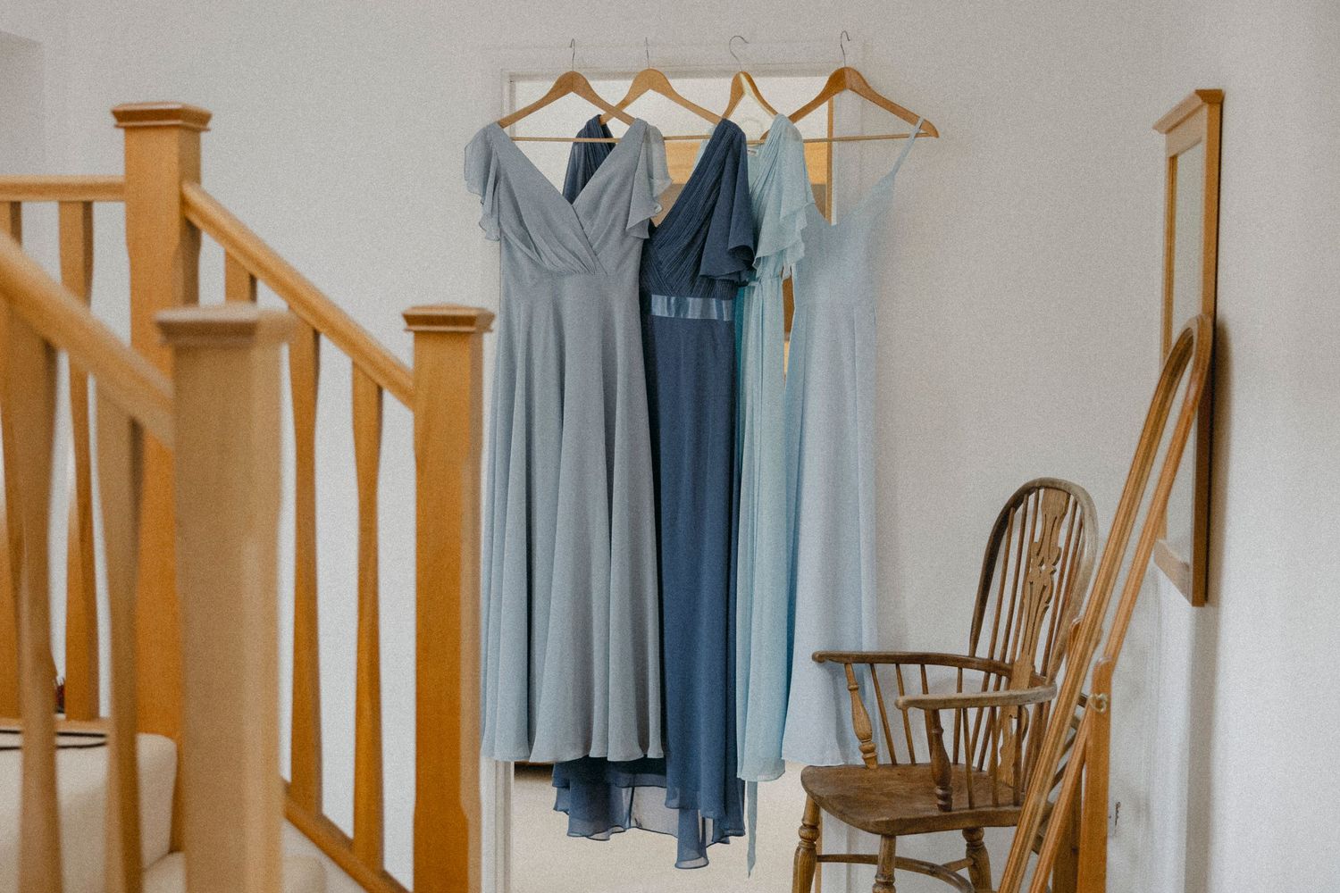 Three blue bridesmaid dresses in varying shades hanging on wooden hangers near a staircase and wooden chair.