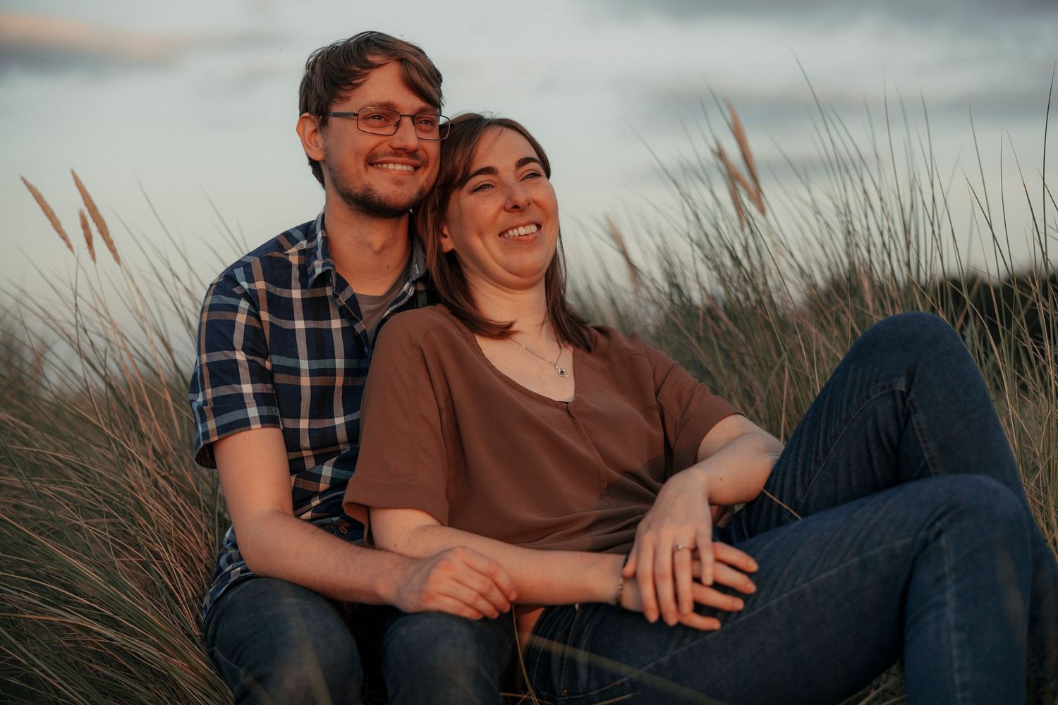 Couple sitting and laughing together in tall grass field during a beautiful sunset.