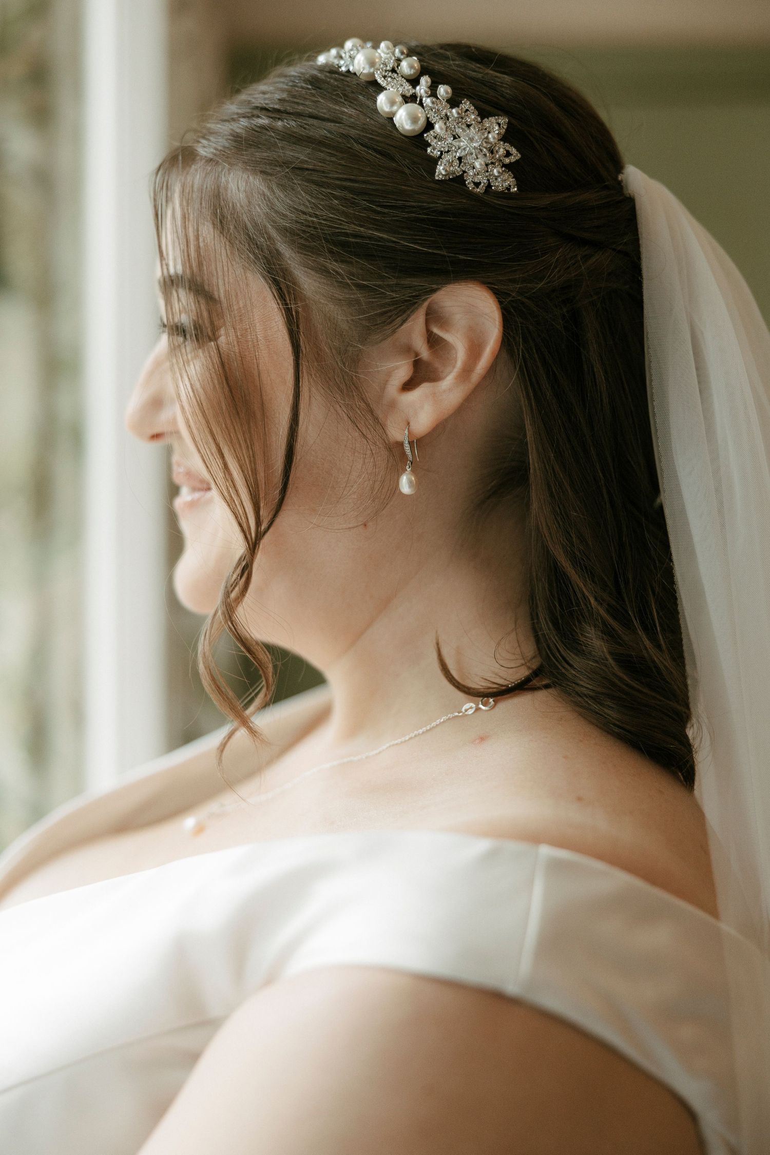 Bride in off-shoulder gown with pearl hair comb and veil, wearing drop earrings and delicate necklace.