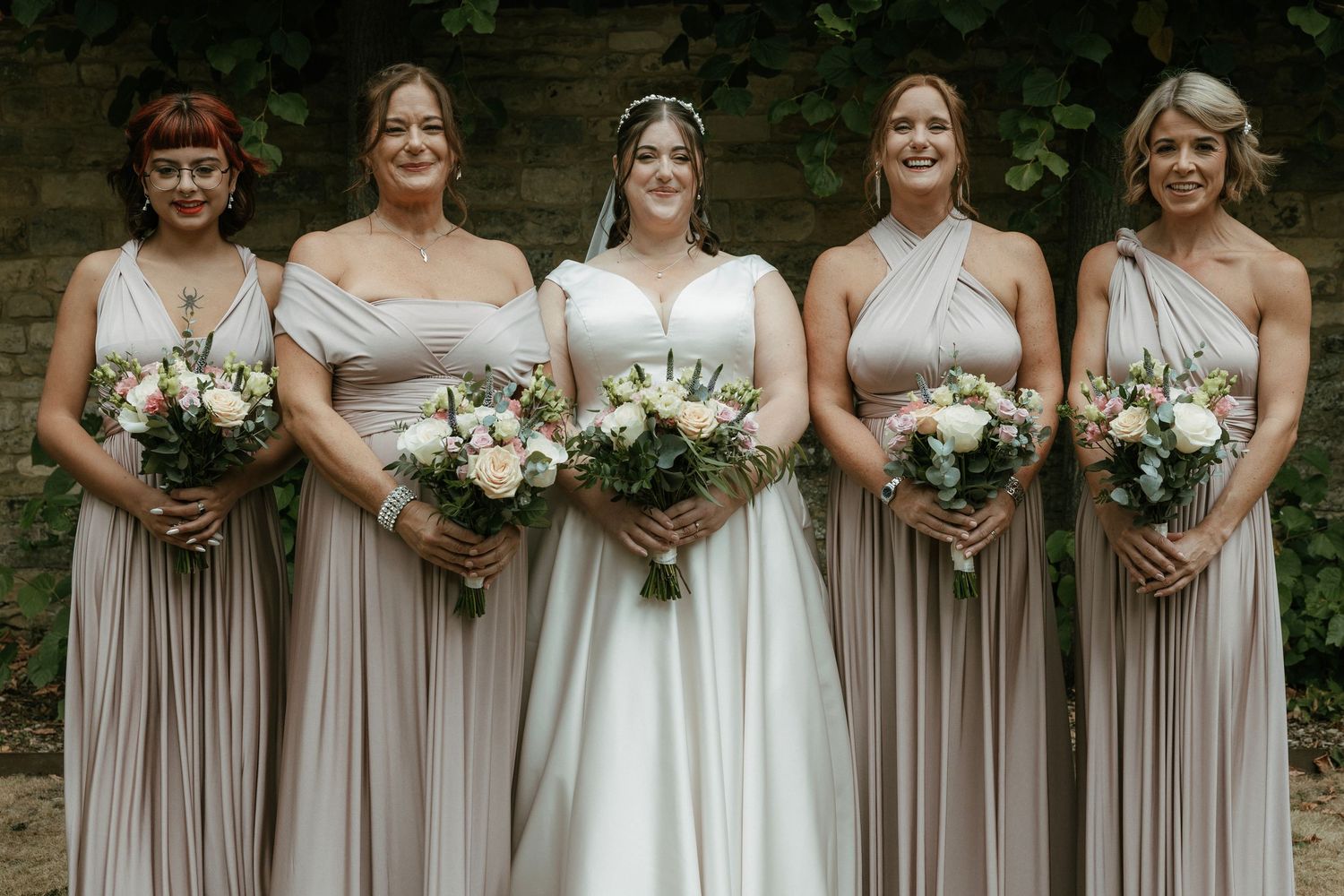Bride in white gown poses with four bridesmaids in taupe multiway dresses, holding bouquets outdoors.