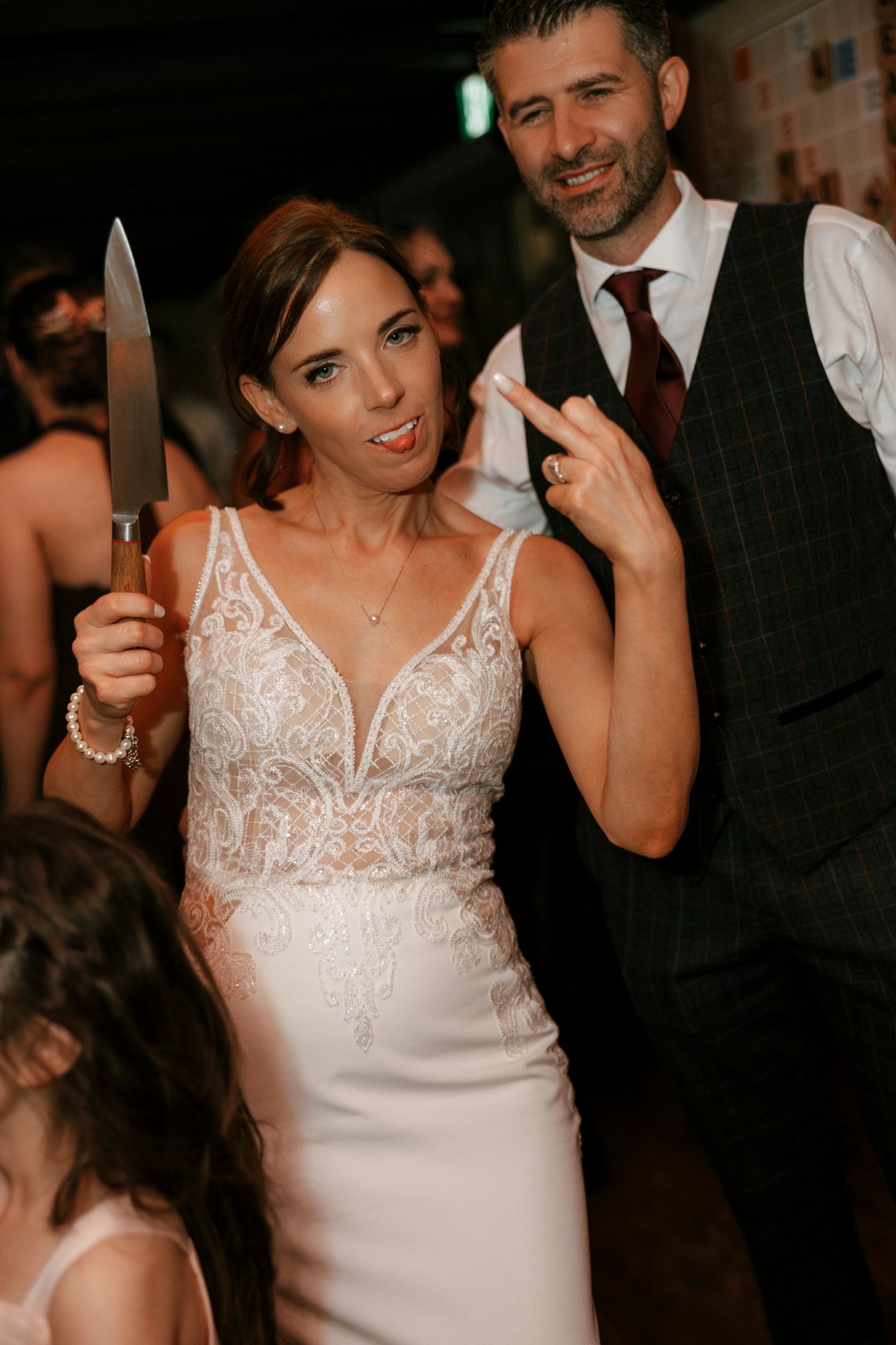 A bride in a white lace wedding dress holds up a large knife at a wedding reception while gesturing enthusiastically.