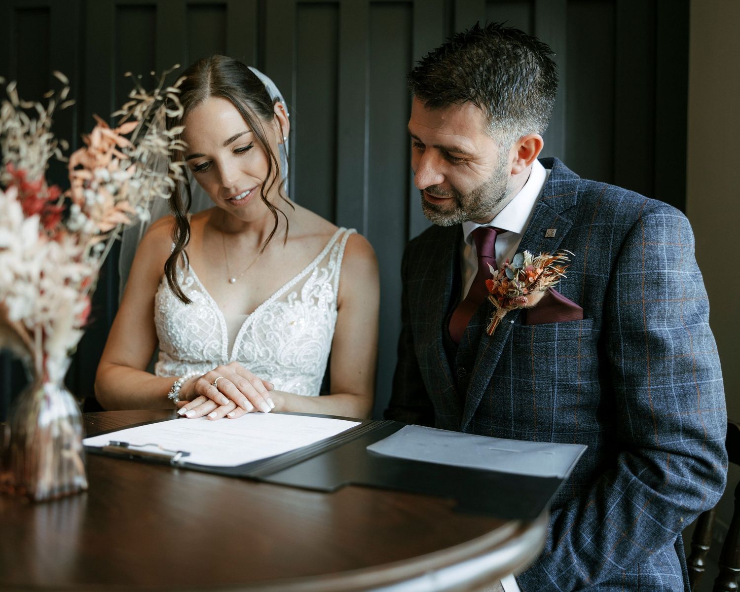 Couple signing wedding documents at a wooden table with floral decor.