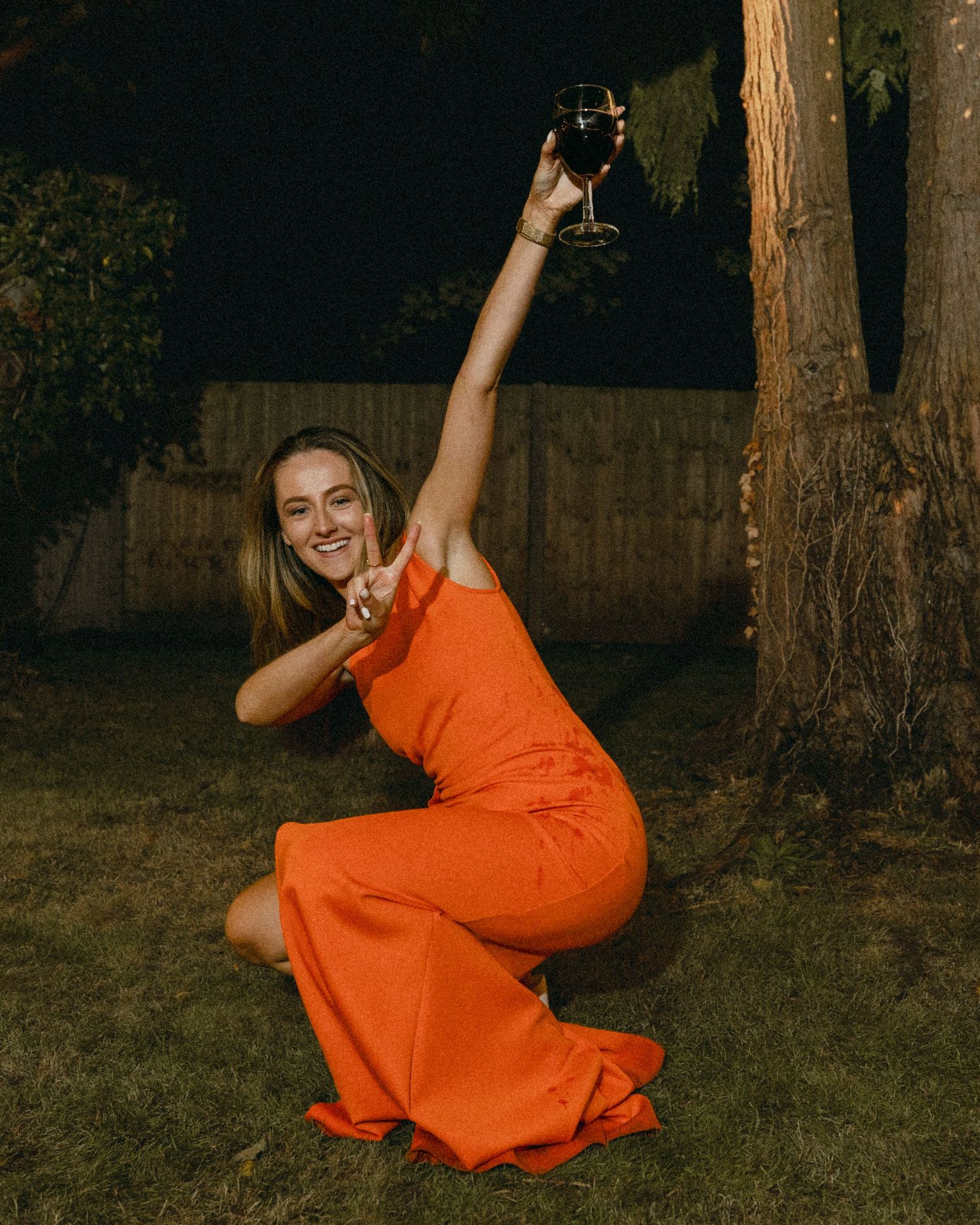 Woman in orange dress sitting on grass, raising a glass of red wine while smiling and making a peace sign.