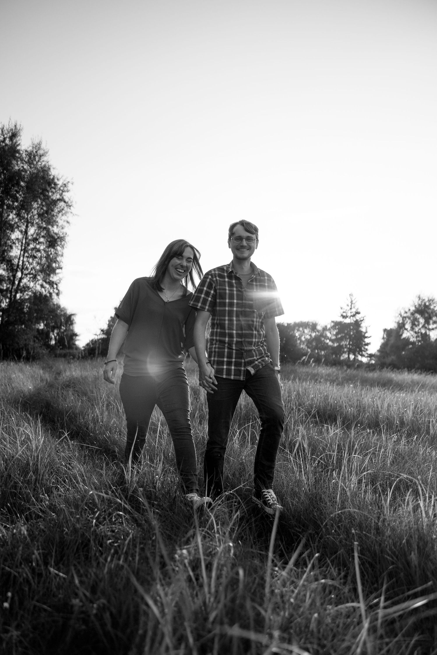 Black and white photo of a happy couple holding hands while walking through a sunlit grassy field at sunset.