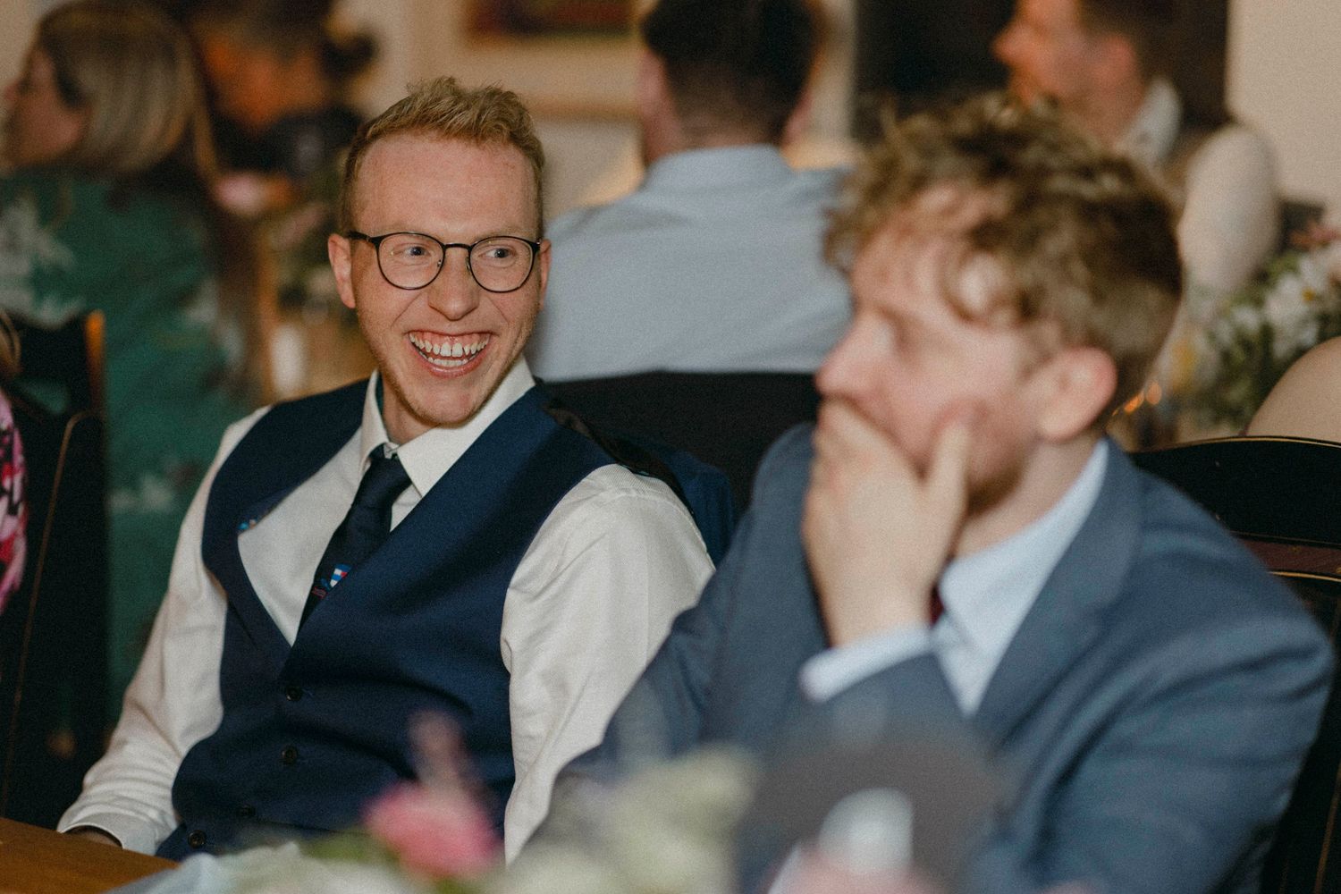 Two men in formal attire smile and laugh at a wedding reception, one wearing glasses and a vest.