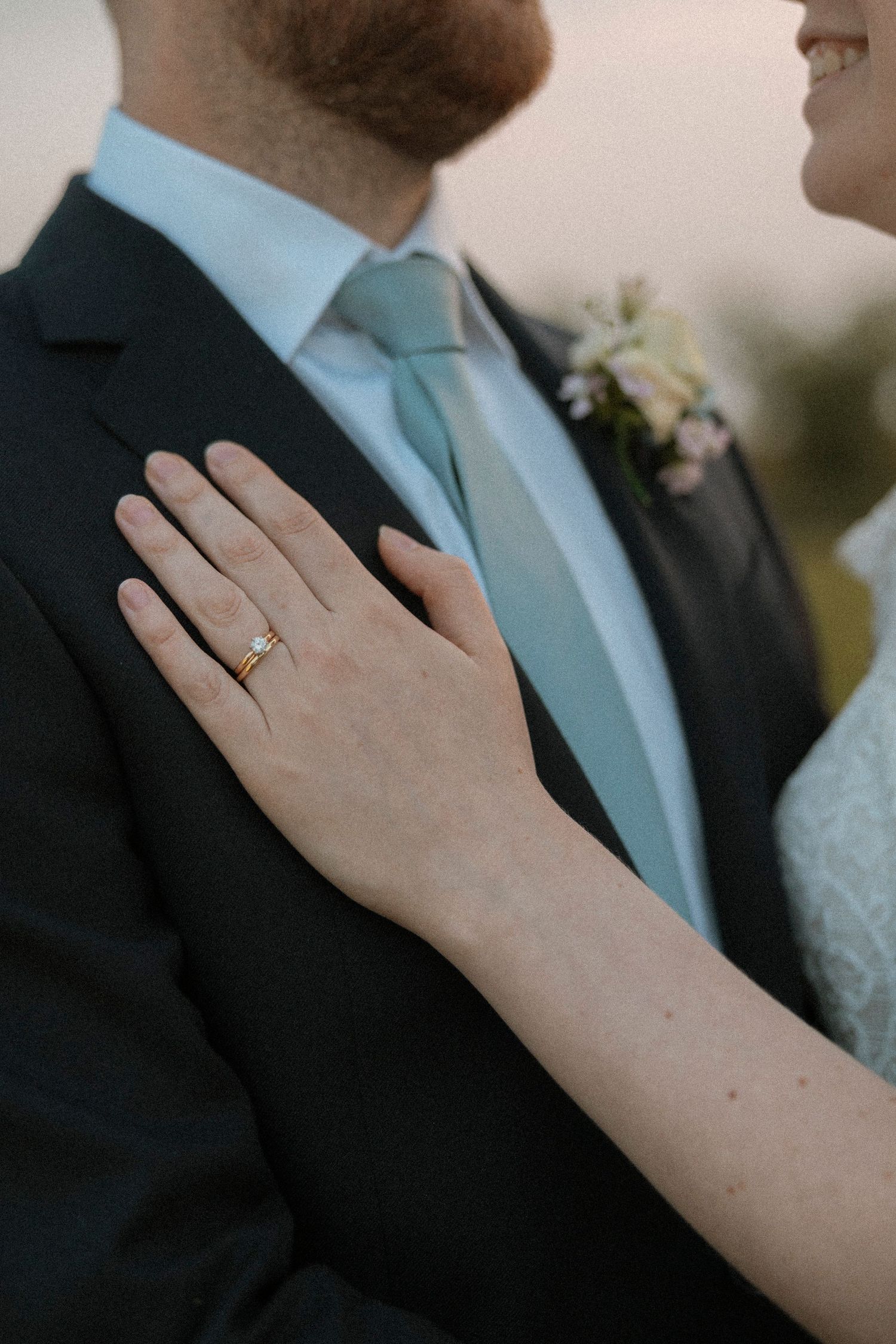 Bride's hand with gold wedding ring resting on groom's dark suit jacket, with teal tie and boutonniere visible.