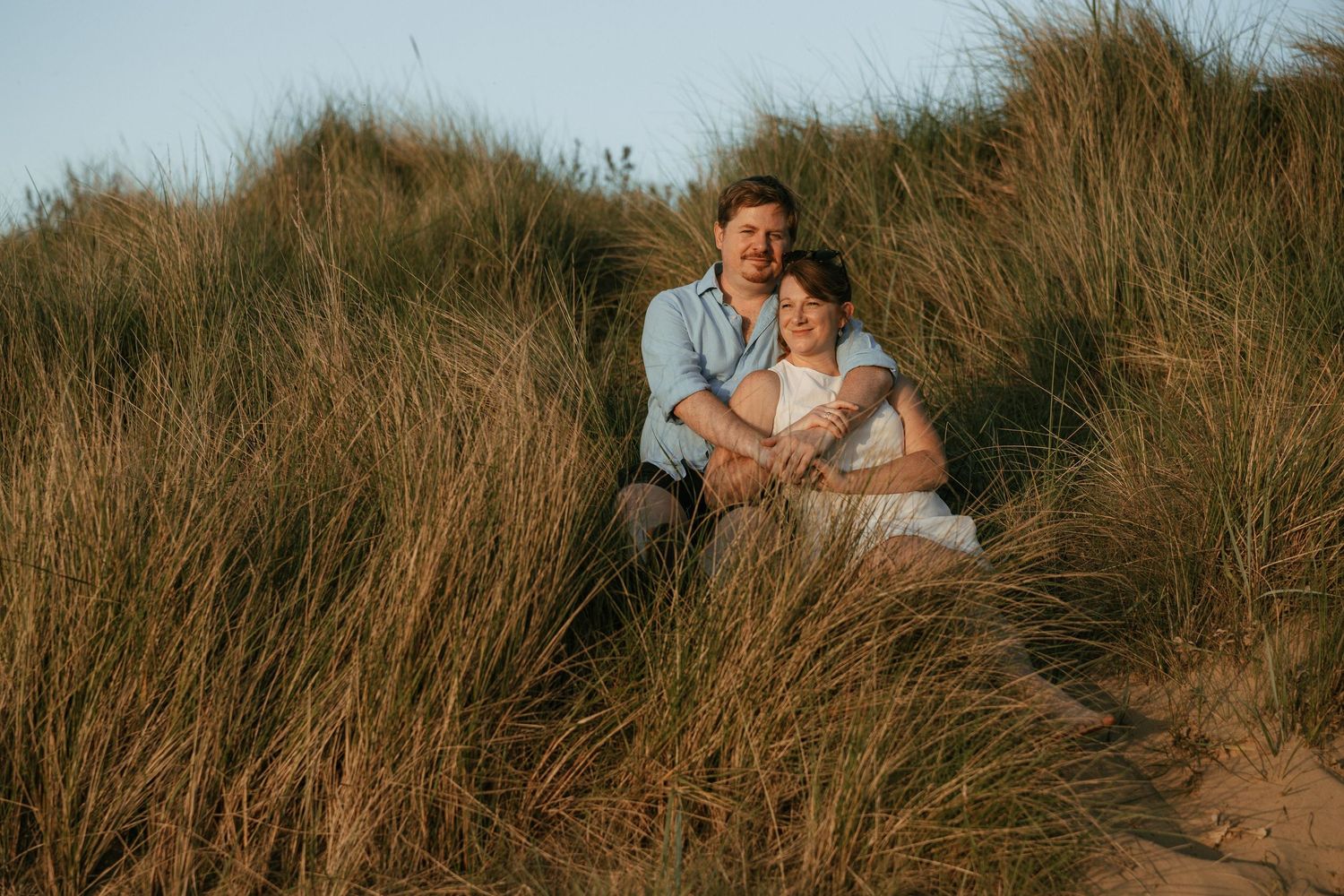 Couple sitting together in golden sand dunes with tall grass, sharing a tender embrace during sunset.