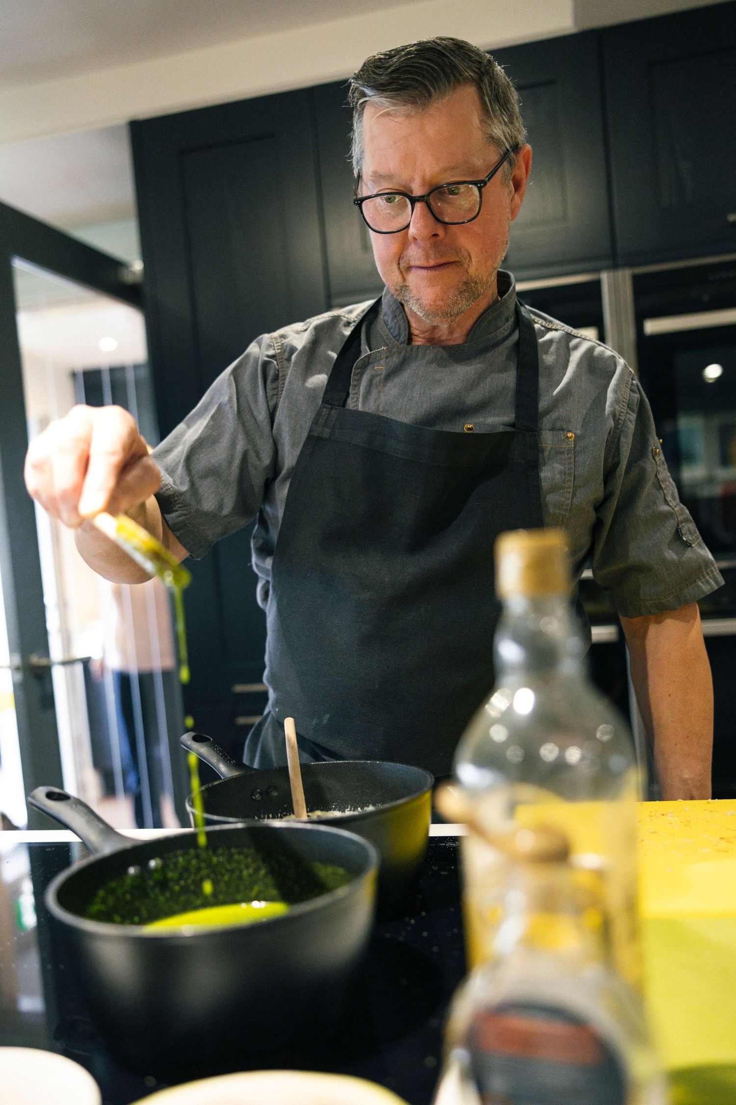 Experienced chef in gray apron and glasses squeezing citrus into a pan of greens on a modern kitchen stovetop.
