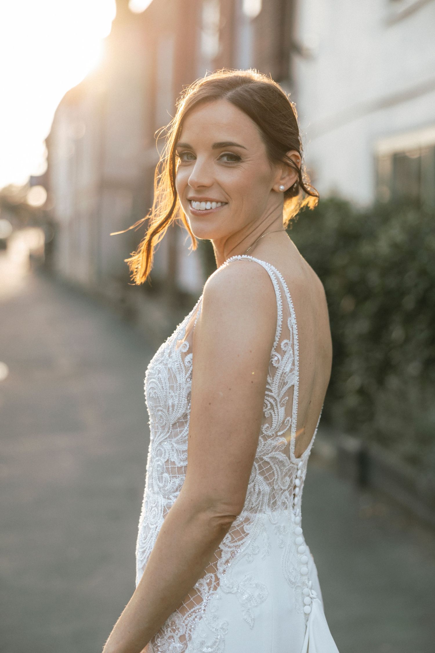 Smiling bride in backless lace wedding dress glowing in golden sunlight on a cobblestone street.