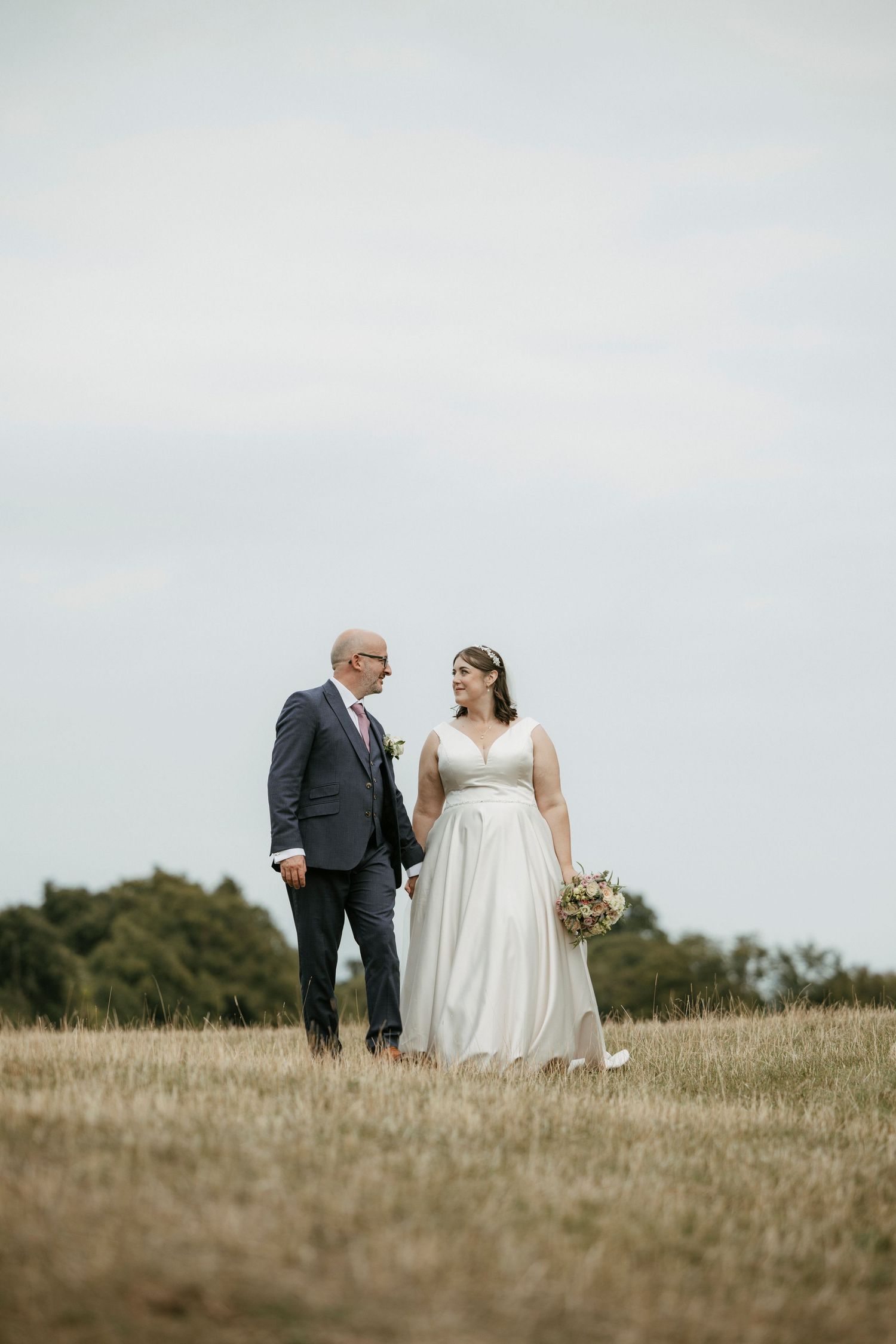 A bride and groom walk hand in hand through a golden field, sharing a loving glance on their wedding day.