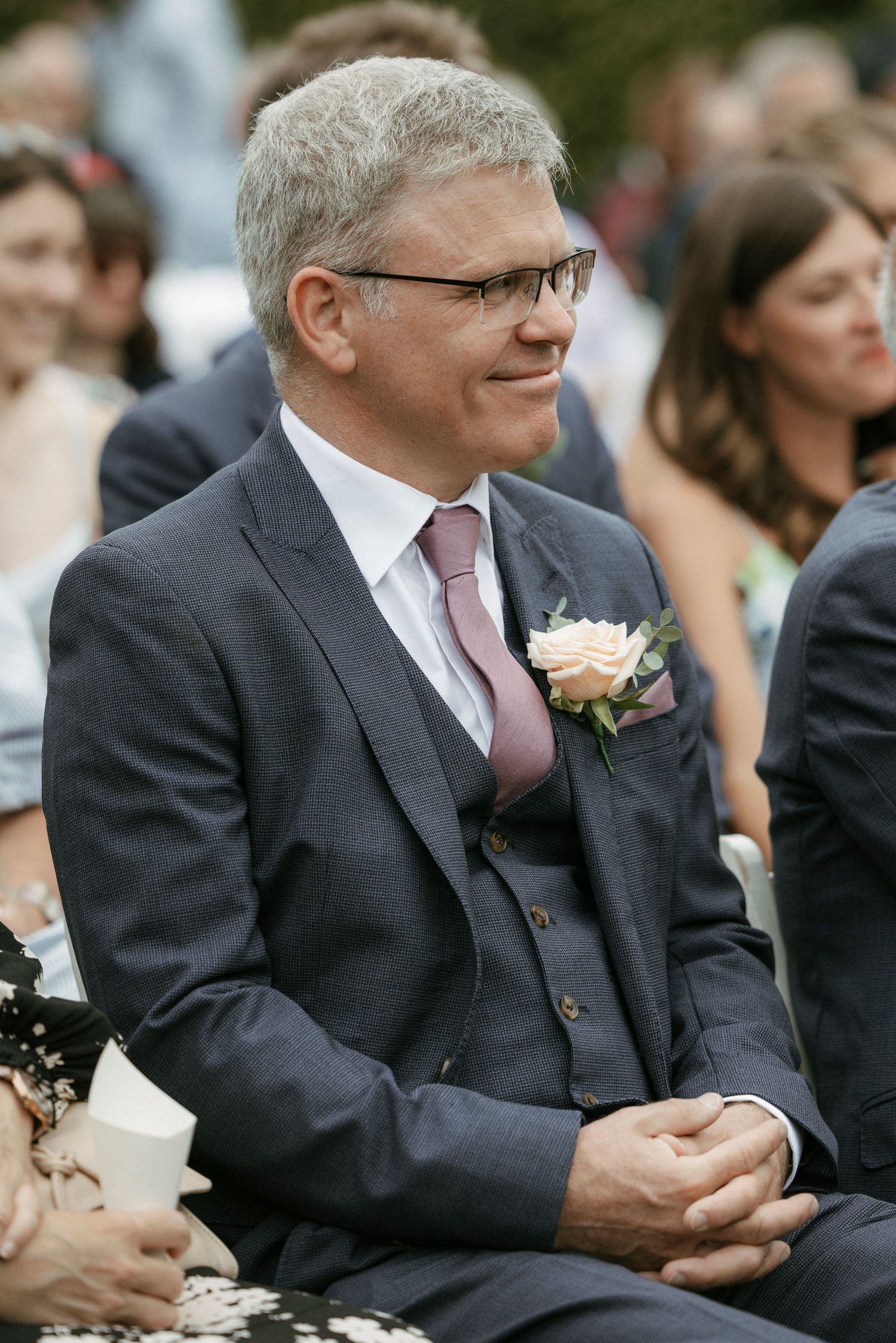 Smiling middle-aged man in gray suit with pink boutonniere and tie seated at outdoor wedding ceremony.