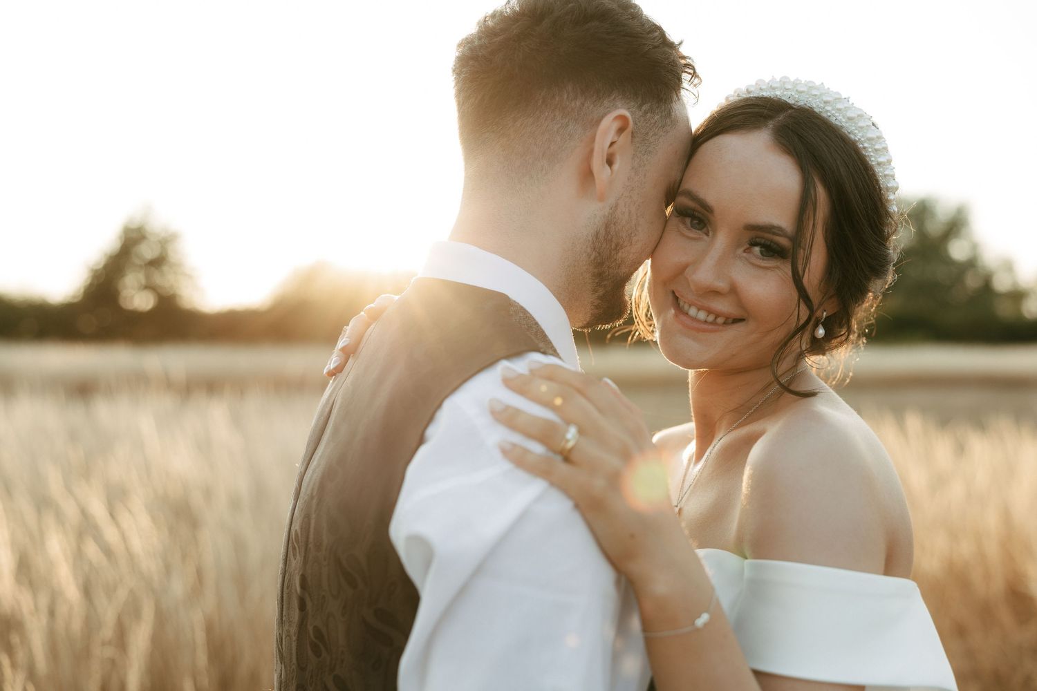 Bride and groom embrace at golden hour in a wheat field, bride smiling warmly wearing a strapless gown and tiara.