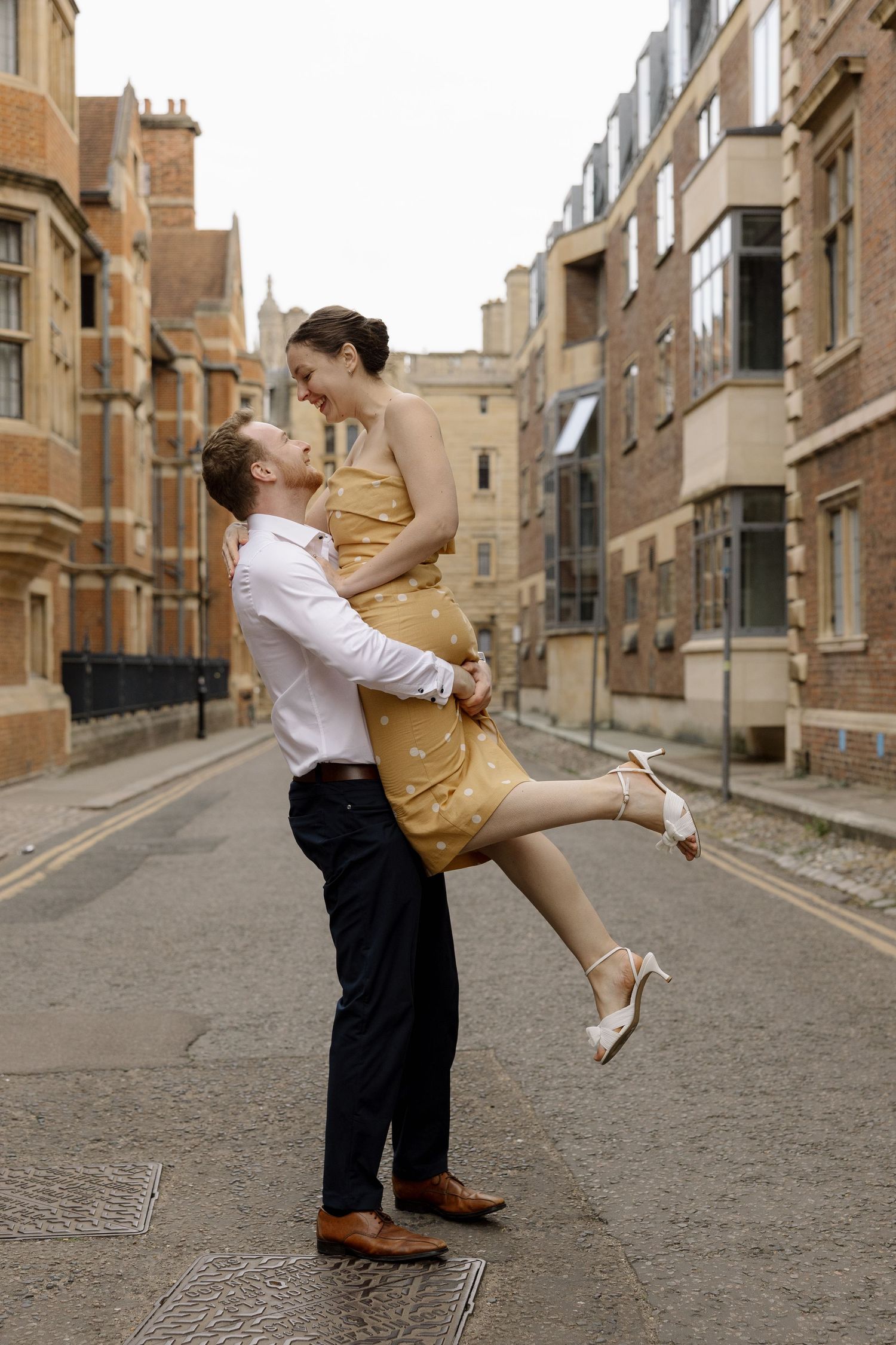 Couple sharing a romantic moment on a cobblestone street, man lifting woman in yellow dress, historic buildings backdrop.