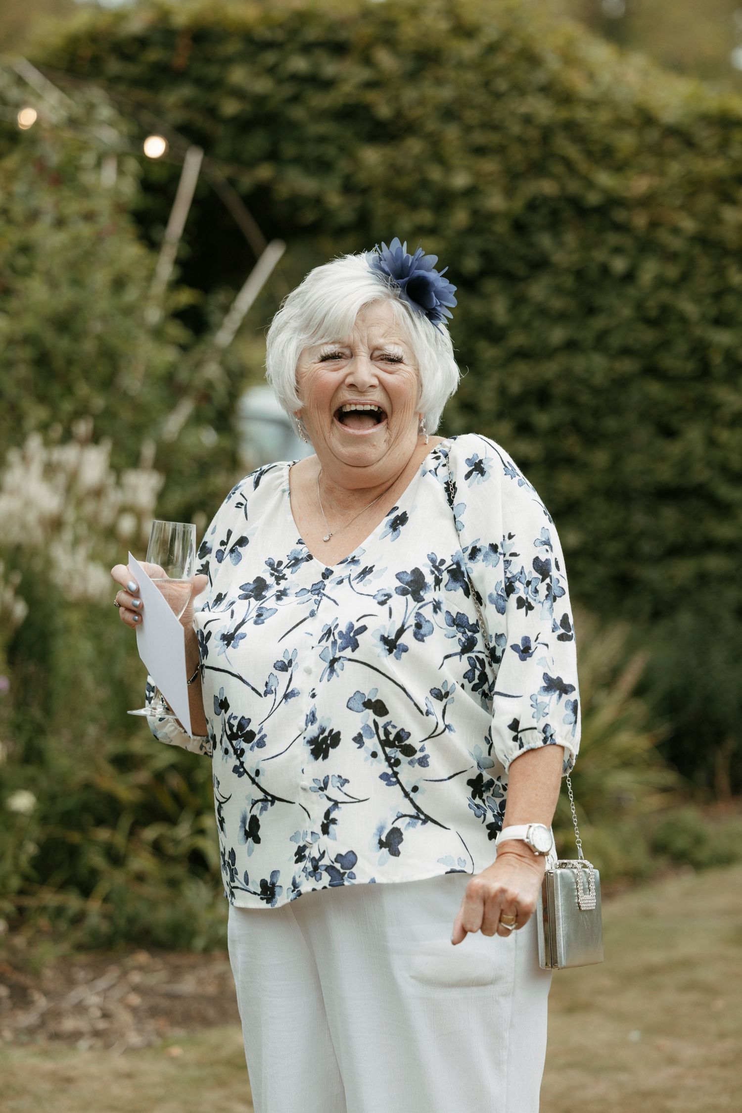 Elderly woman laughing joyfully at outdoor garden party, wearing floral blouse, white pants, blue hair accessory, holding champagne glass.