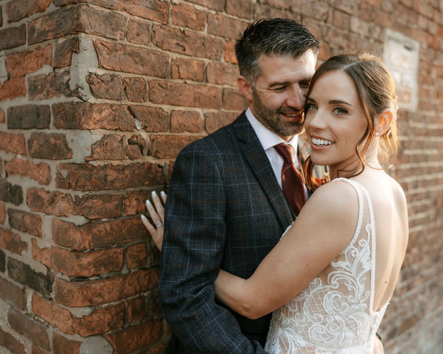 Bride and groom embrace against rustic brick wall, groom in plaid suit with red tie, bride in lace wedding gown smiling.
