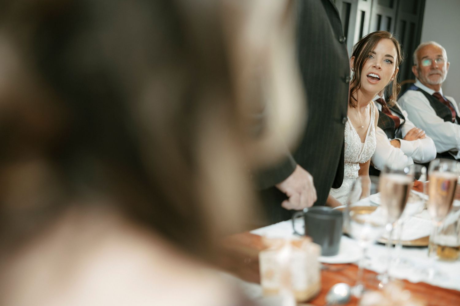 Joyful woman laughing at a wedding reception dinner table with guests and champagne glasses in background.