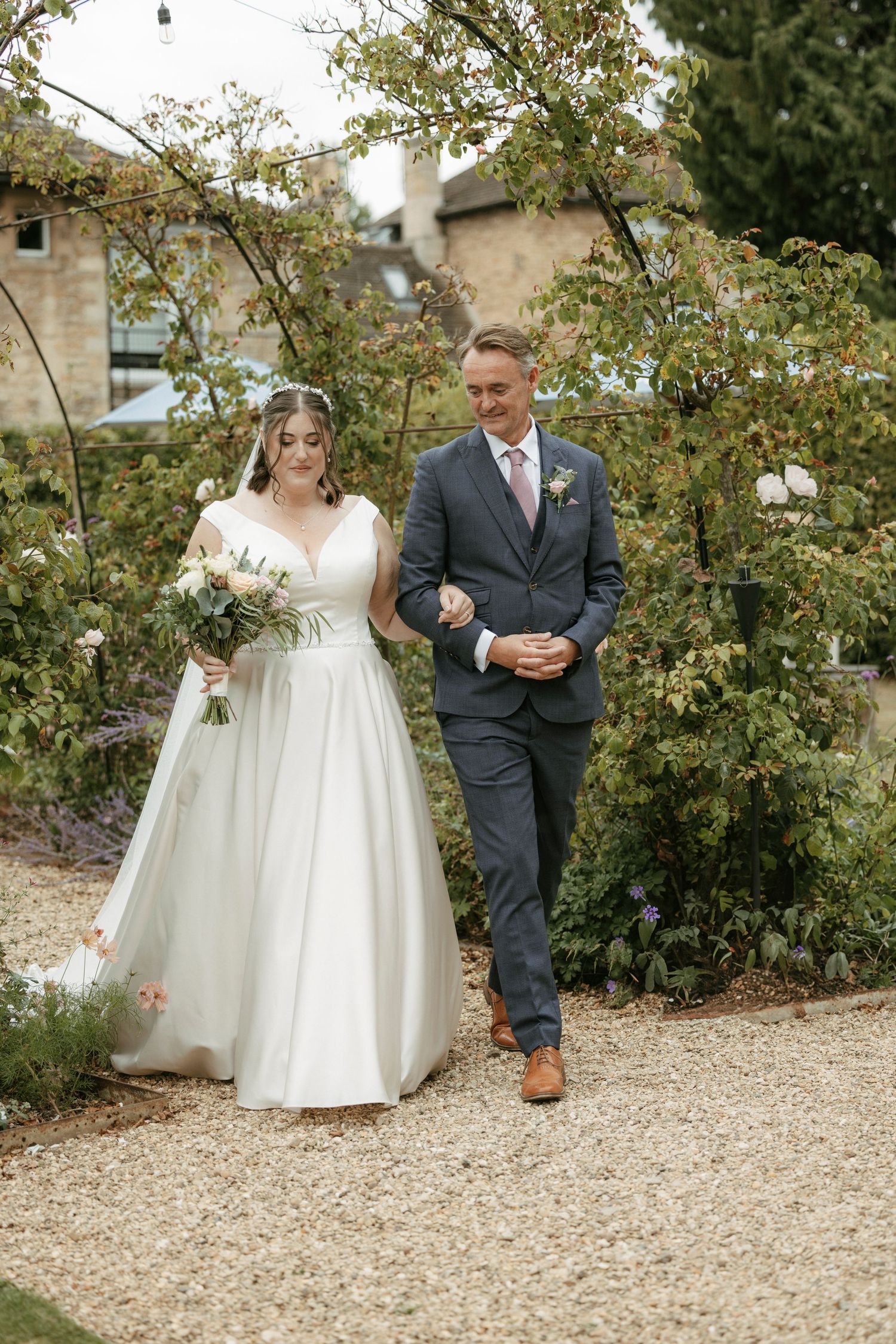 Bride holding bouquet walks arm-in-arm with escort along garden path with stone building backdrop.