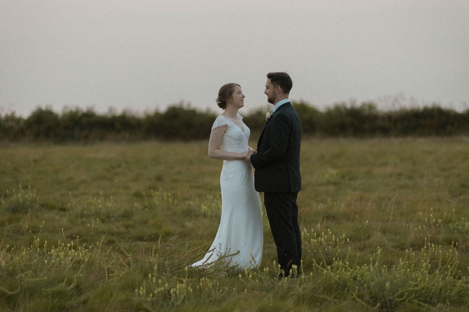 A bride and groom stand facing each other in an open green field on a cloudy day, sharing a romantic wedding moment.