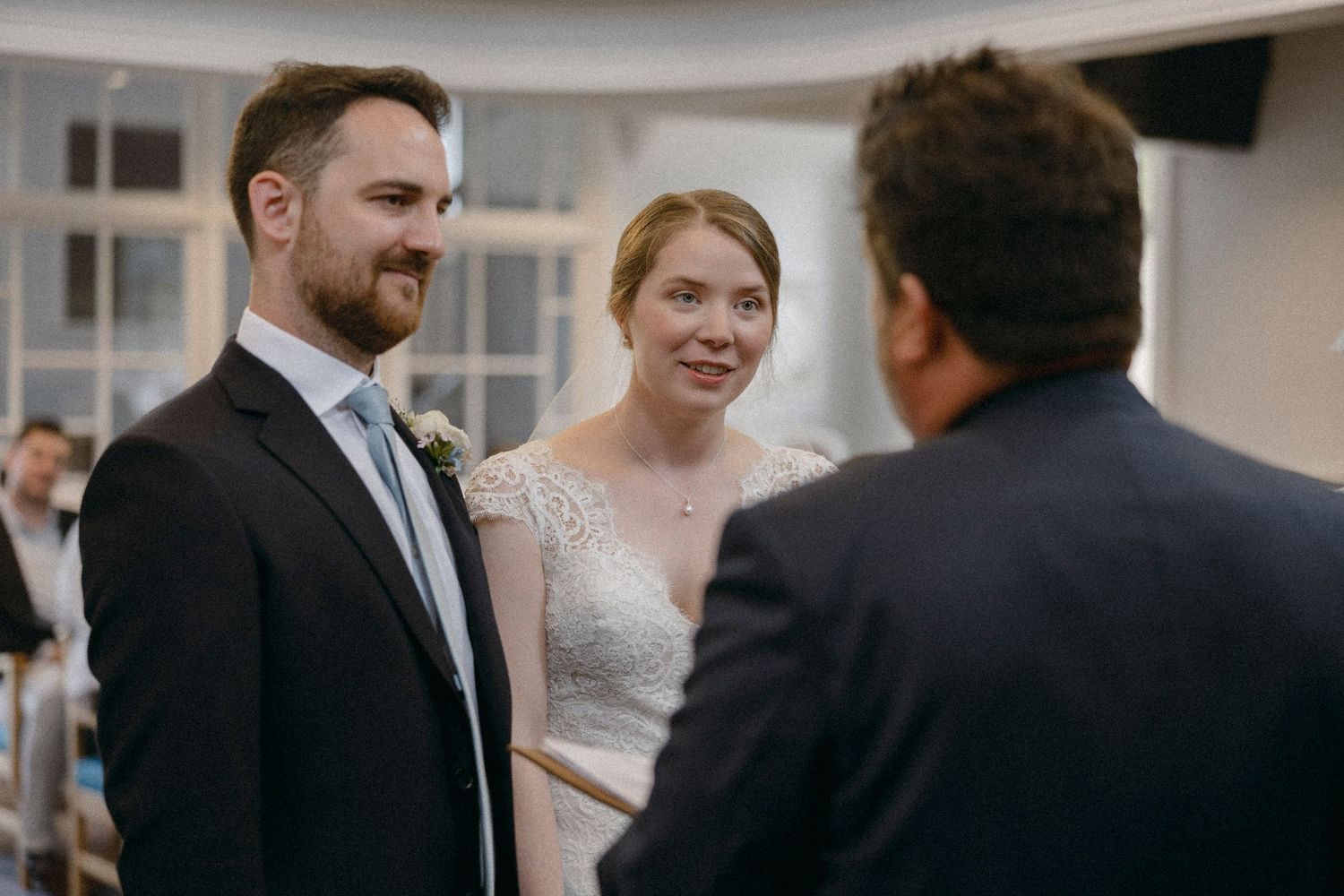 A bride and groom smile during their wedding ceremony while speaking with an officiant in an elegant indoor venue.