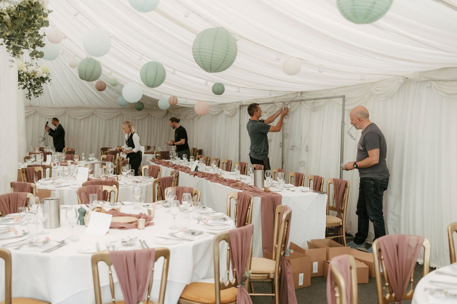 Elegant wedding marquee being decorated with sage green paper lanterns, pink chairs, and white round tables set for a reception.