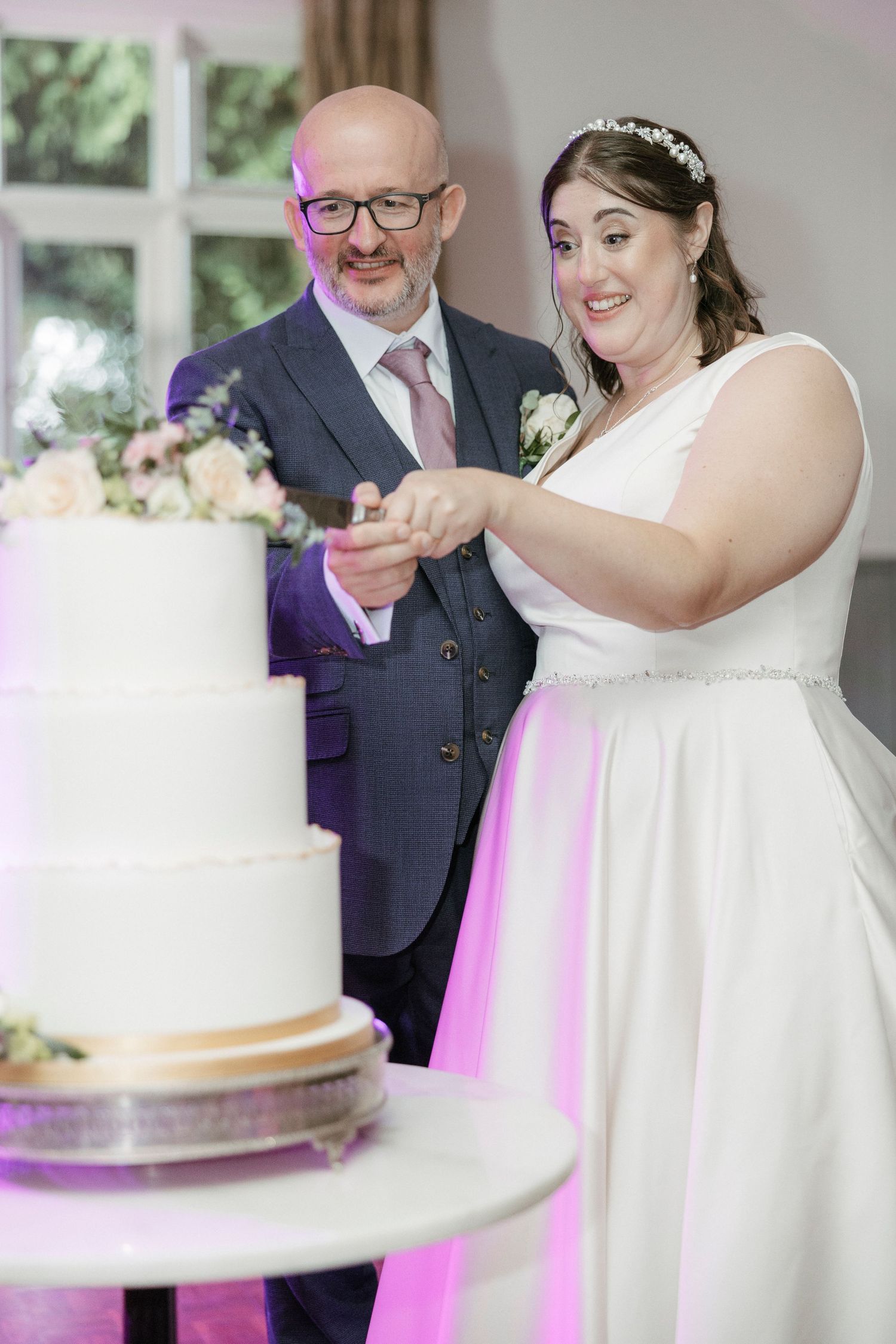 Bride and groom smiling while cutting white three-tier wedding cake together.