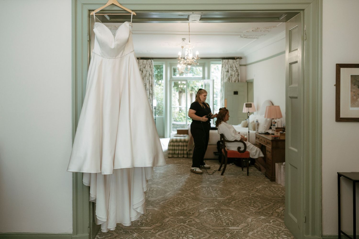 White wedding gown hanging in doorway while bride gets hair done in elegant room.