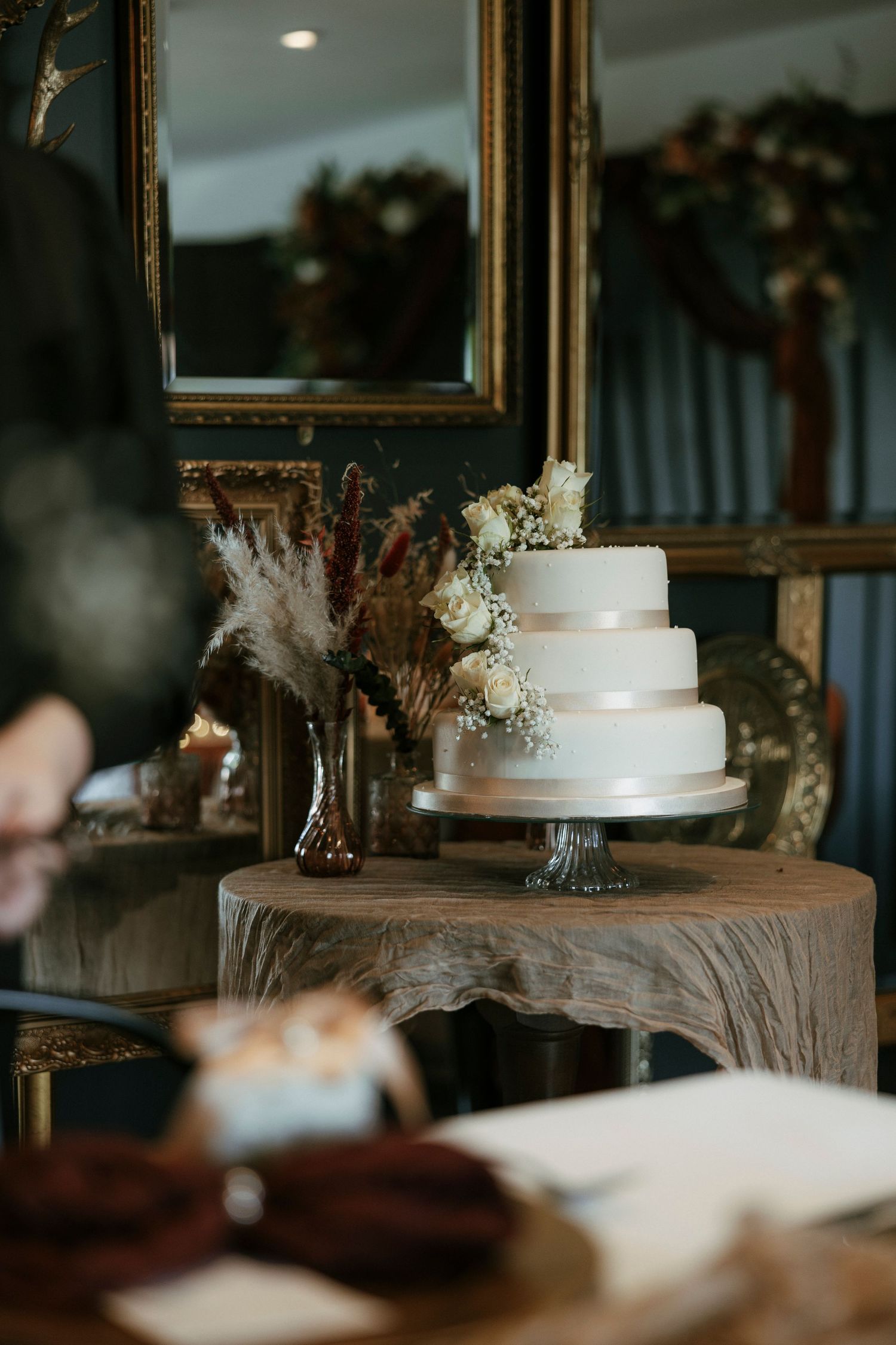 Elegant three-tiered white wedding cake displayed on rustic wood stand with dried florals and ornate gold mirror backdrop.