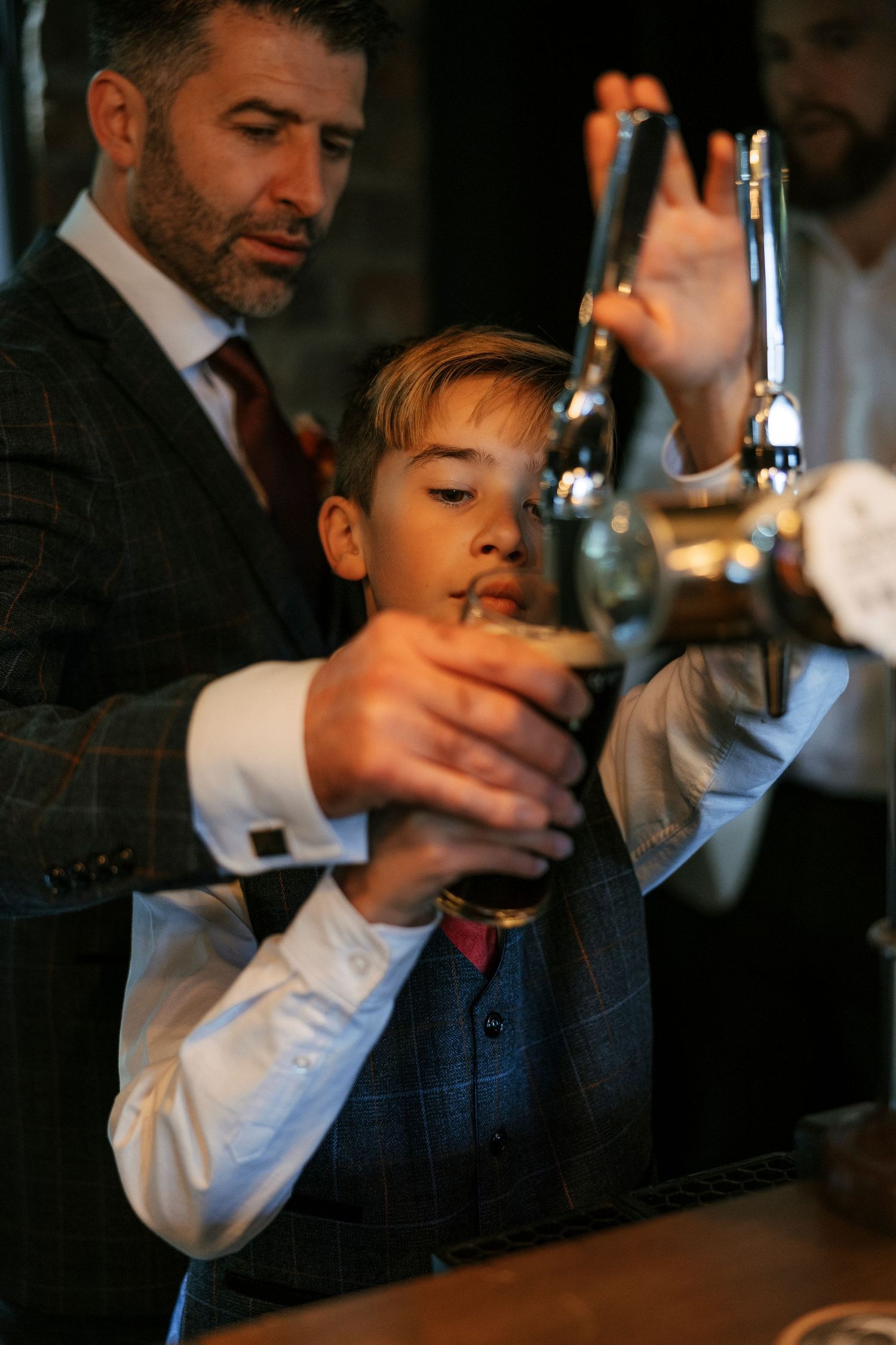 Adult pouring a beverage at bar with youth looking on in formal attire.