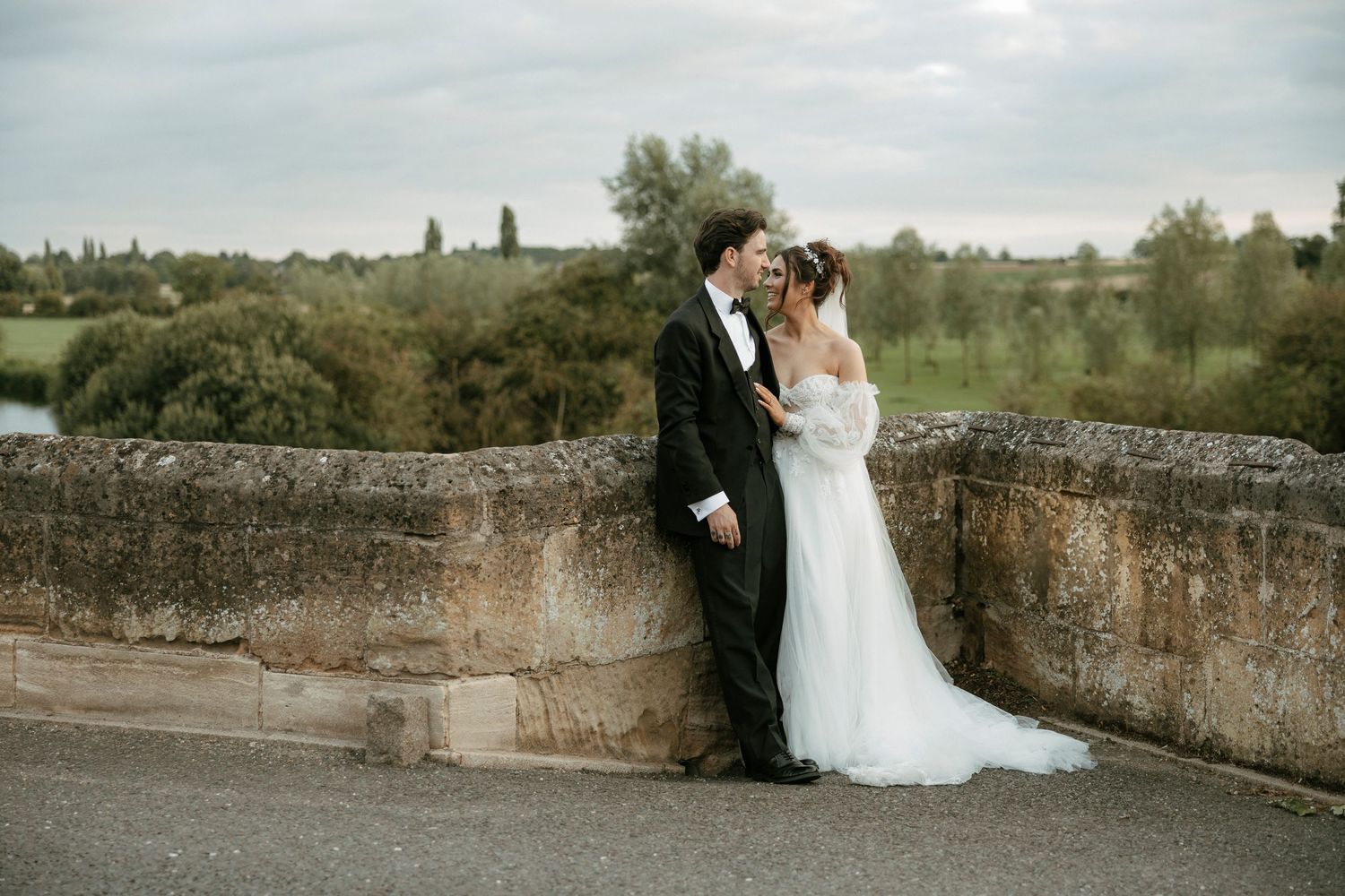 A romantic wedding couple poses on a rustic stone bridge in a scenic countryside setting.