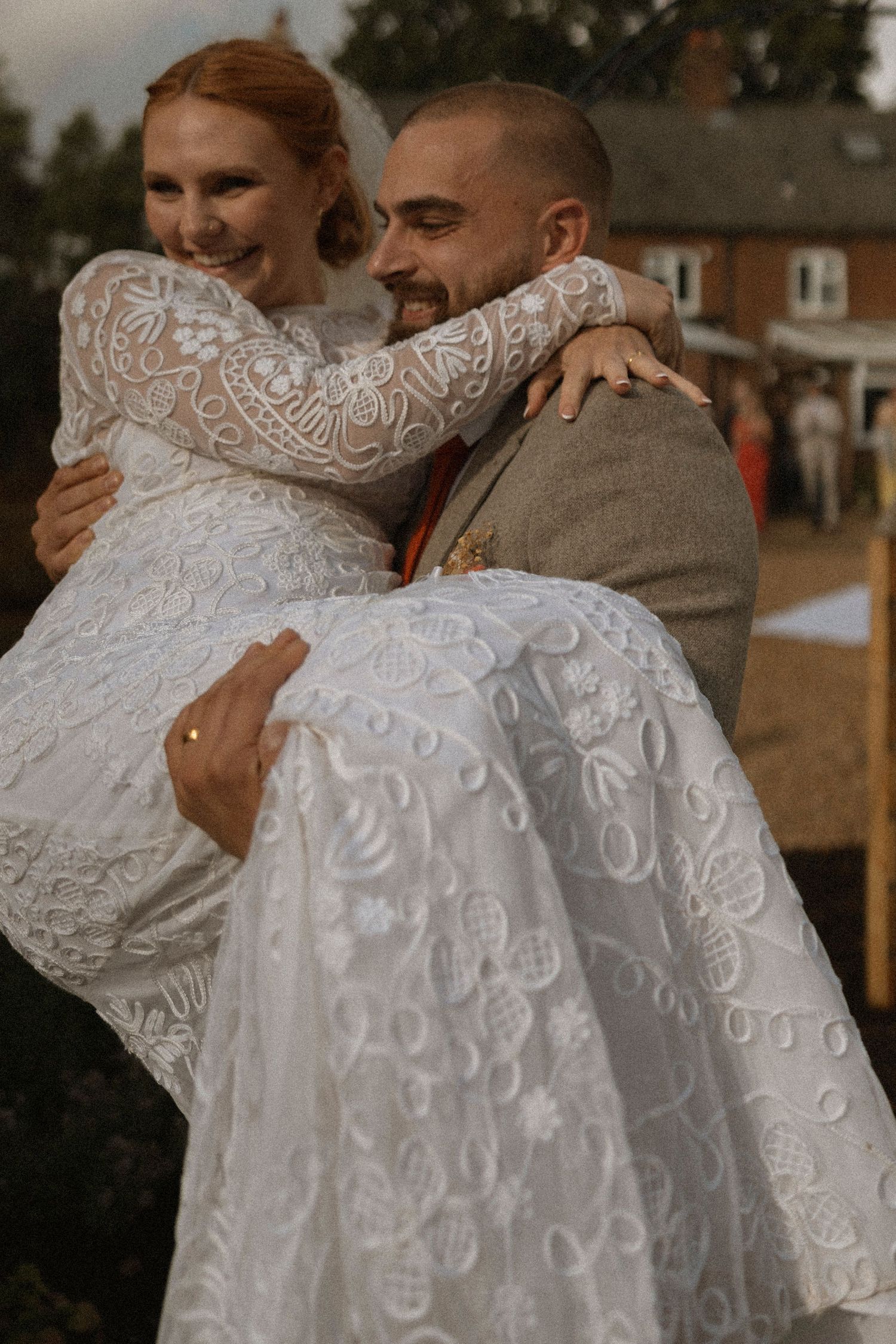 Groom carrying bride in elegant lace wedding dress, both smiling joyfully outdoors at a countryside wedding venue.