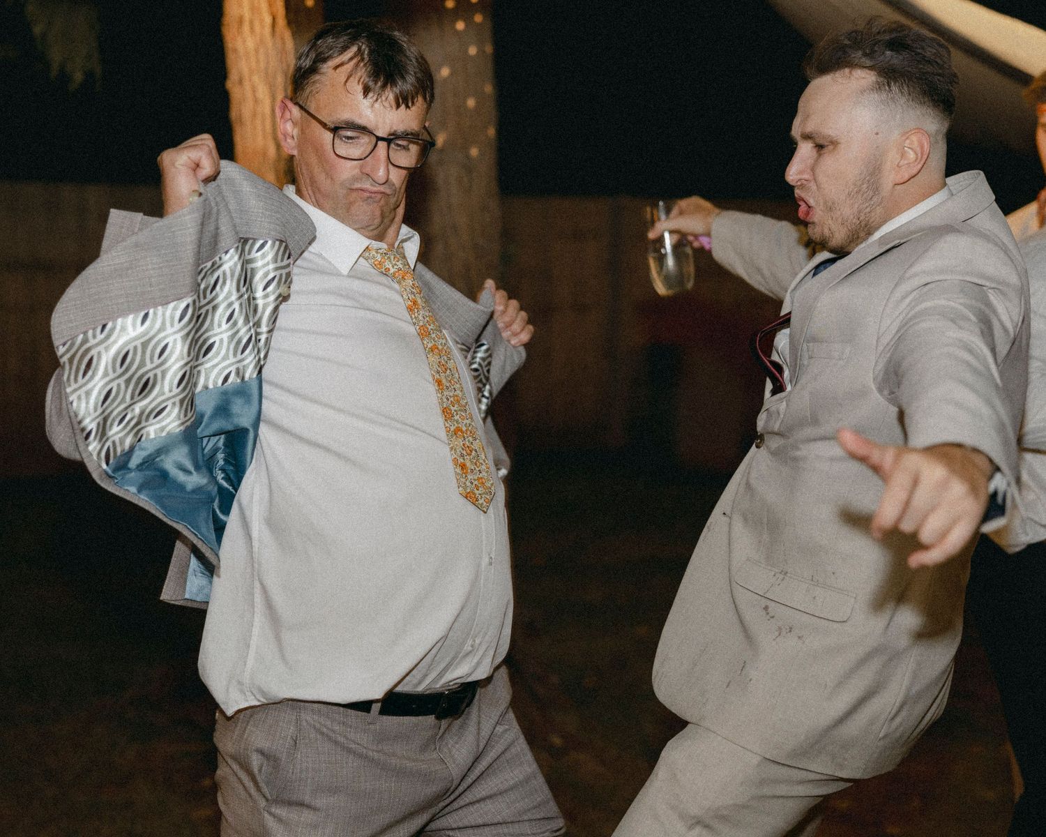 Two men in suits dancing energetically at a wedding reception party at night.
