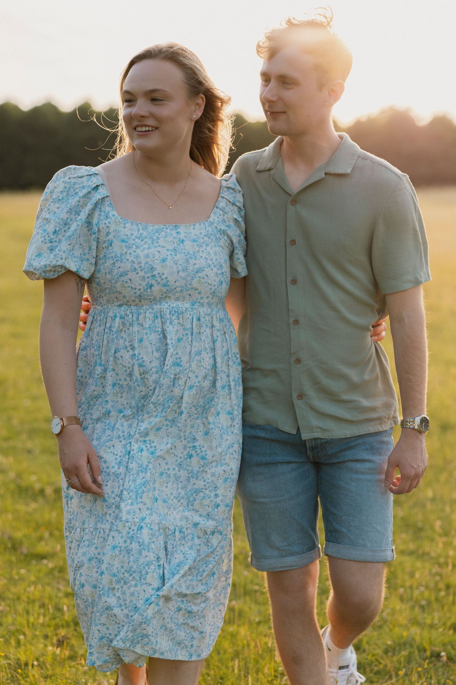 Happy couple walking together in sunny green field, woman in blue floral dress, man in sage shirt.