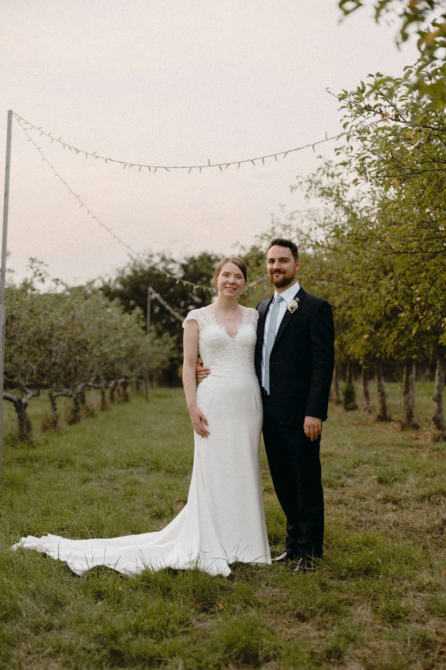 Bride and groom posing in an orchard with string lights, bride in white gown, groom in dark suit.