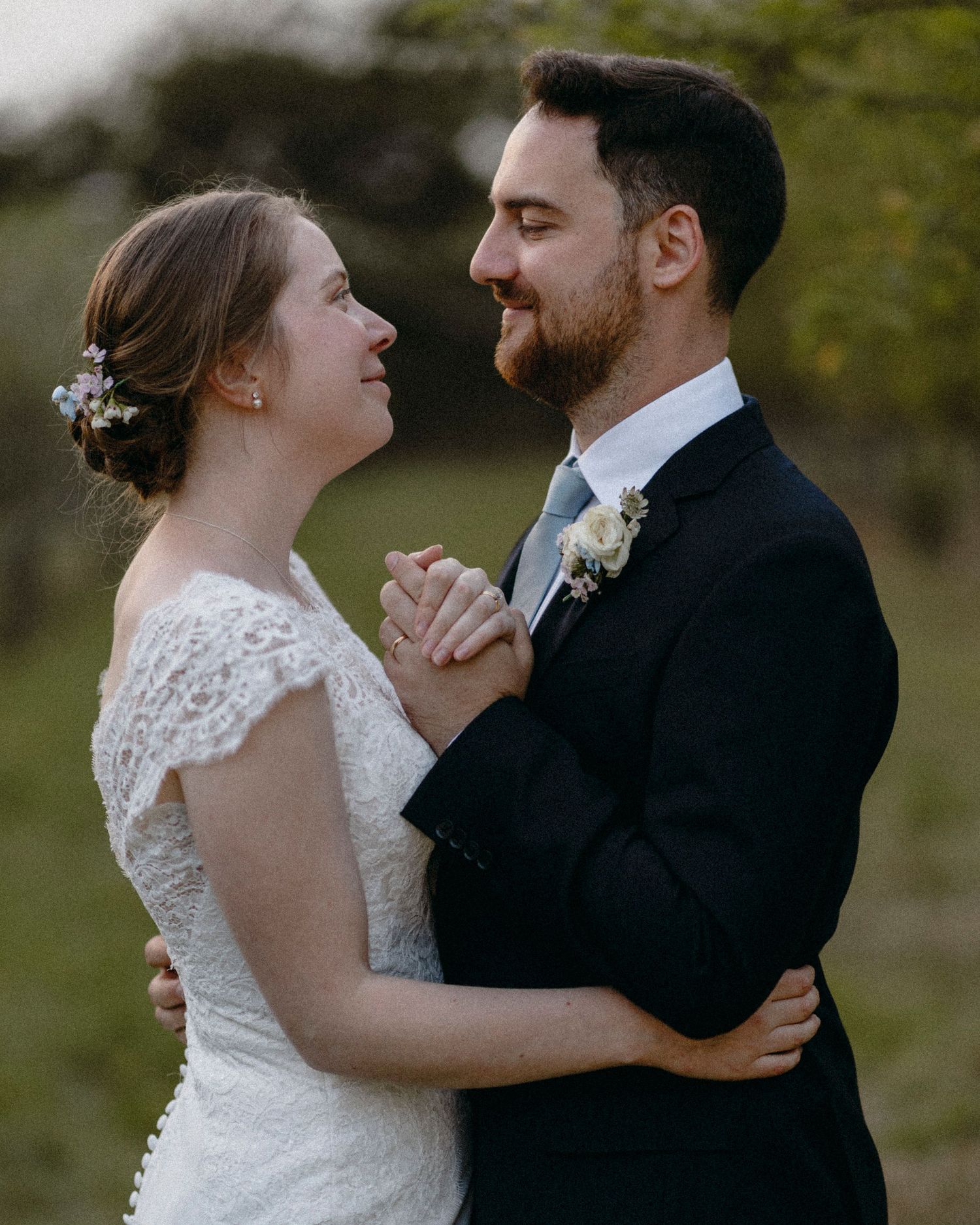 A bride and groom share a tender moment, holding hands and gazing at each other in wedding attire outdoors.