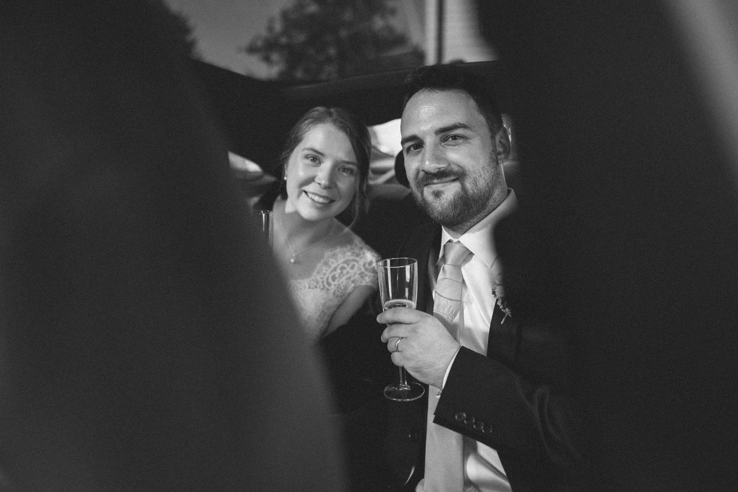 Black and white photo of a smiling couple at a wedding, man holding champagne glass, woman in elegant dress beside him.