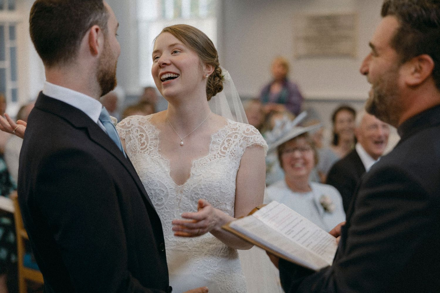 Bride laughing joyfully during wedding ceremony while exchanging vows with groom before officiant in church.