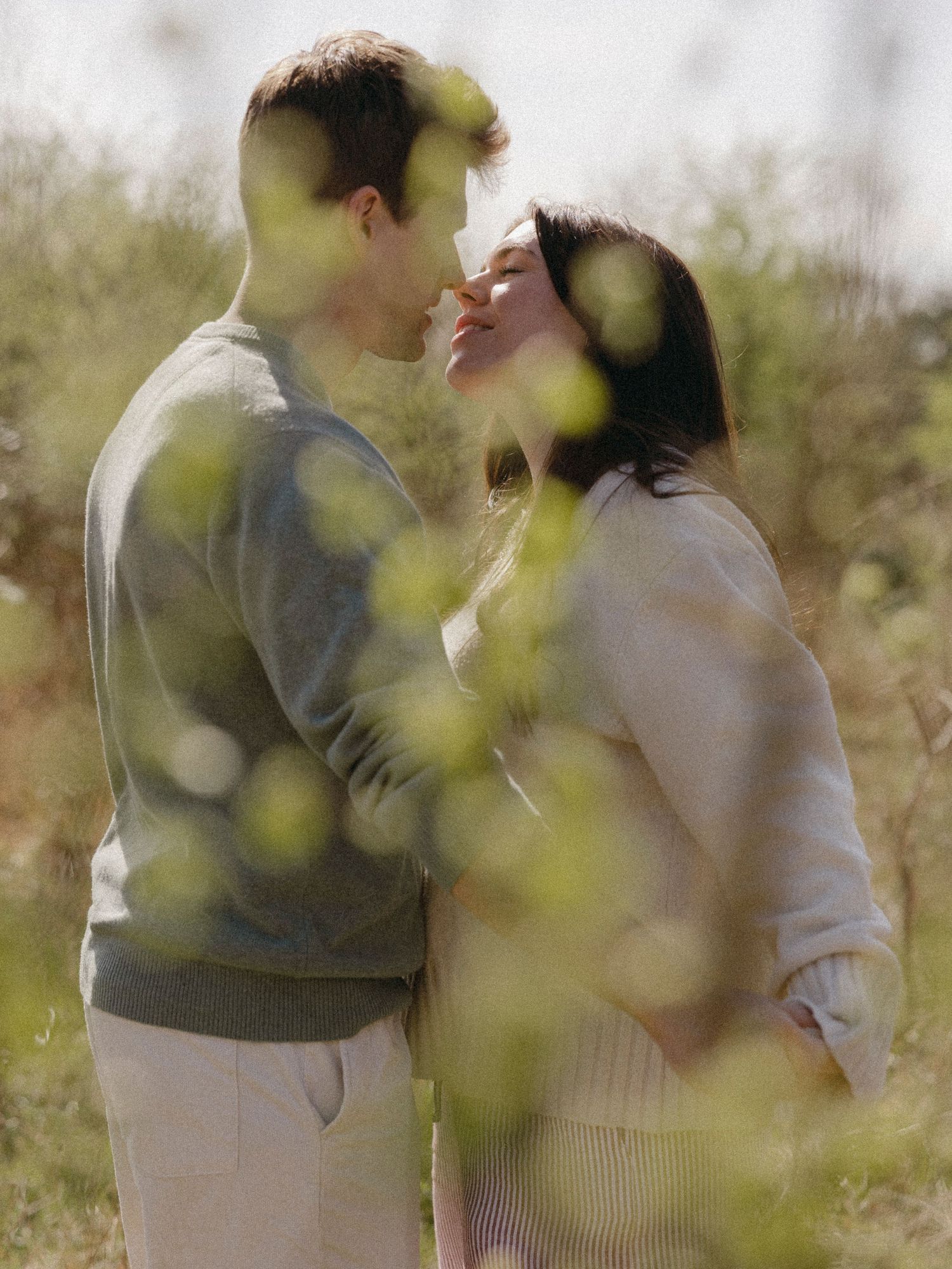 Couple sharing a tender moment outdoors, surrounded by soft bokeh greenery in warm natural light.