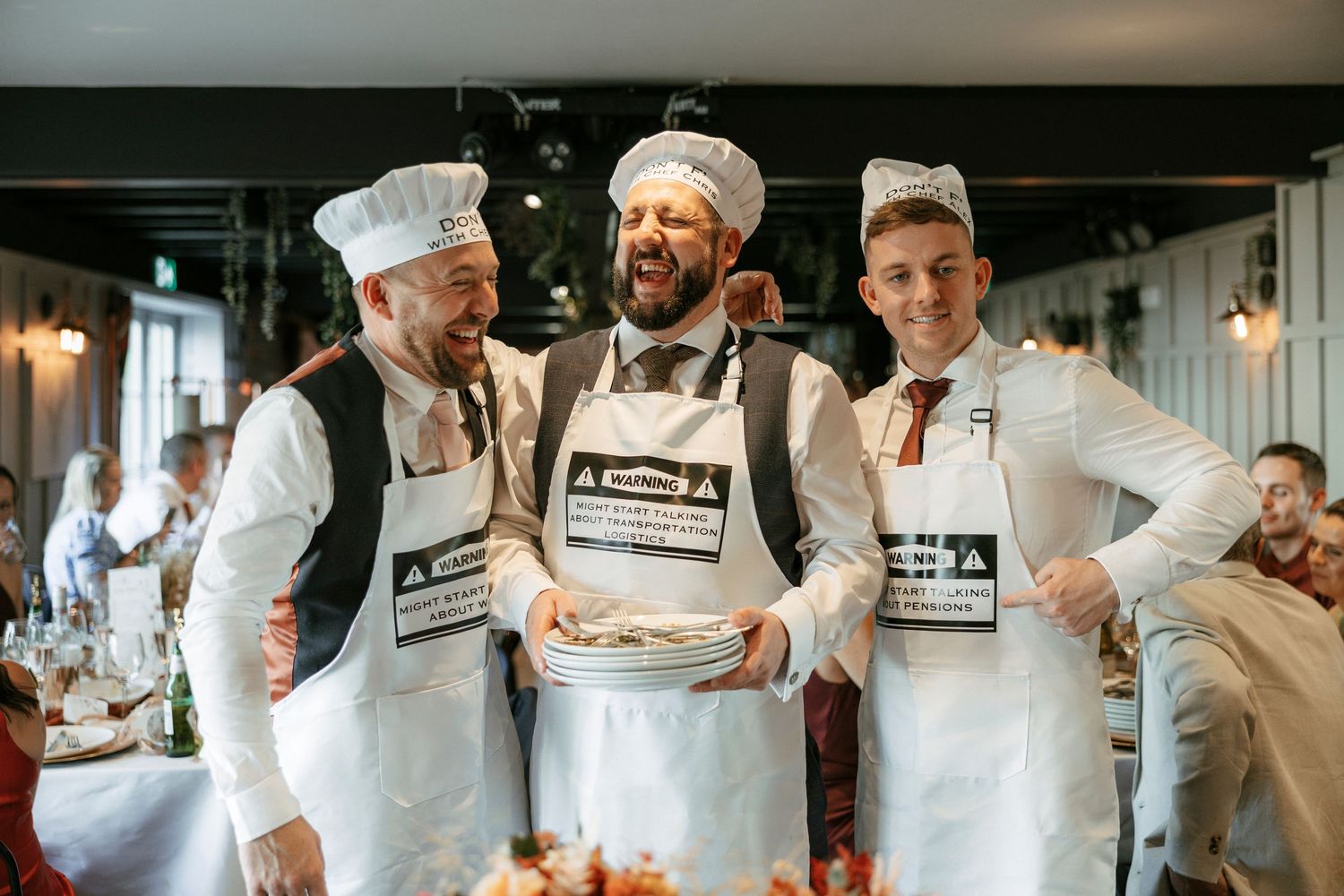 Three men in chef hats and white aprons smile while holding plates in a festive restaurant setting.