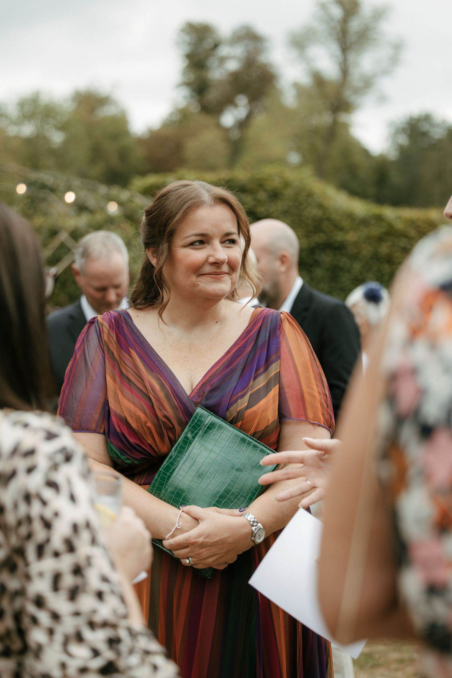 Woman in colorful striped dress holding green book at outdoor wedding reception, smiling warmly at guests.
