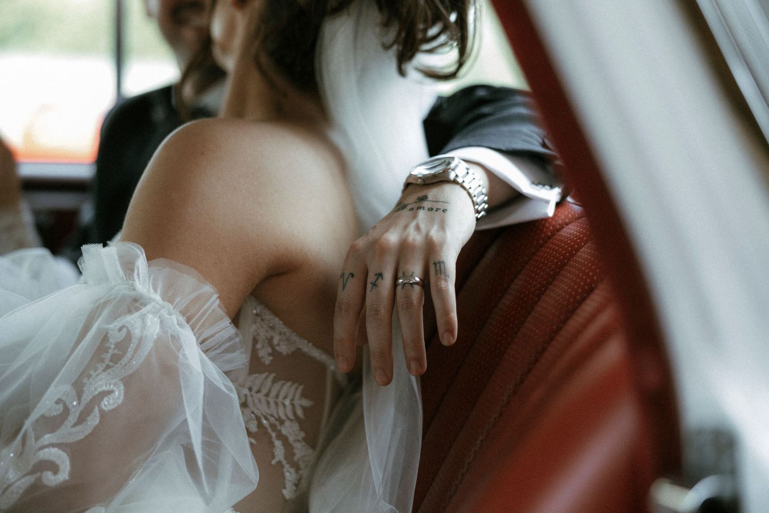 Bride in lace wedding dress showing diamond ring while sitting on red leather car seat beside groom.