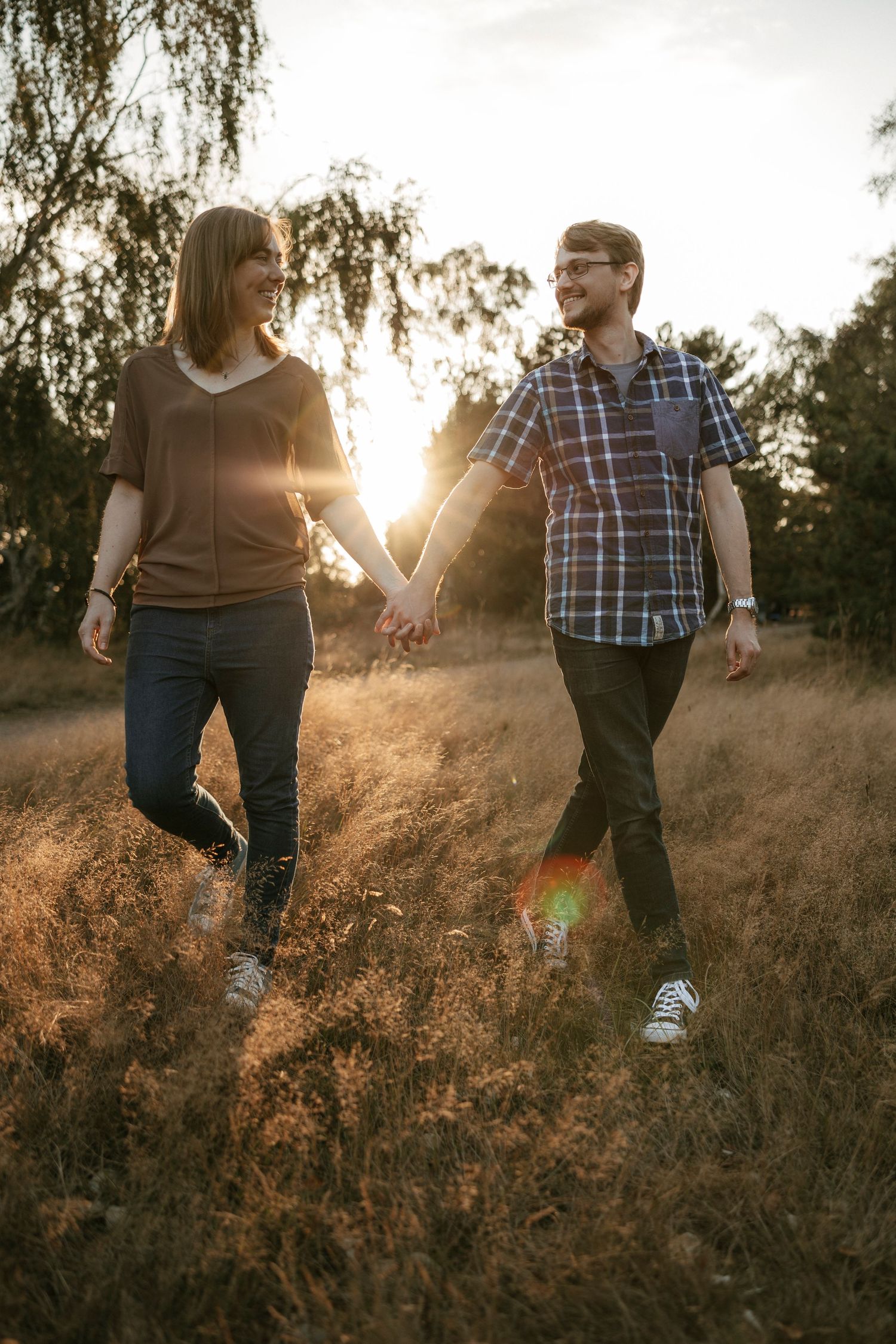 Couple holding hands walking through golden field at sunset, smiling at each other in warm backlit light.
